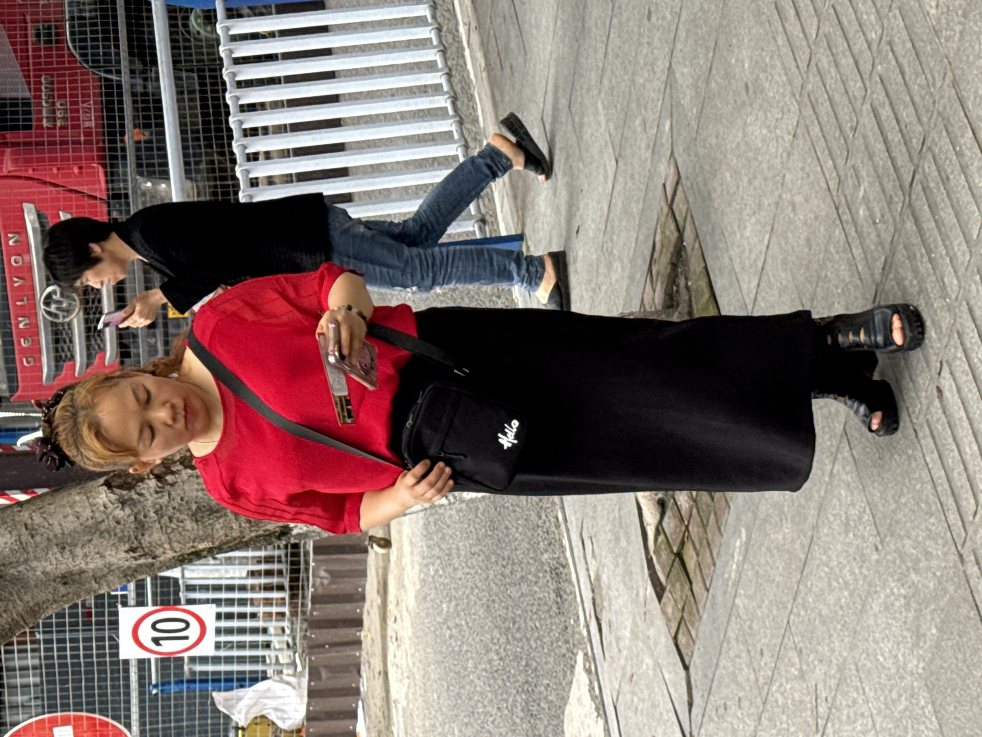 A medium-angle outdoor shot shows two people walking along a sidewalk in what appears to be an urban environment in Jiangjin, China. A woman in a bright red long-sleeved shirt and a long black skirt is in the foreground, facing the viewer's left and slightly downward. She carries a black crossbody bag with "Hello" written on it in white, and she holds a phone in her right hand. She wears black sandals.
Behind her, a person with short dark hair is seen walking in the same direction. They wear a black shirt and blue jeans, and are looking at a phone in their hands. They wear black sandals.
In the background, there is a tree trunk and a metal fence. A red vehicle is visible behind the fence, and a road with a speed limit sign "10" is present. The ground is paved with concrete tiles. The lighting suggests it is daytime.