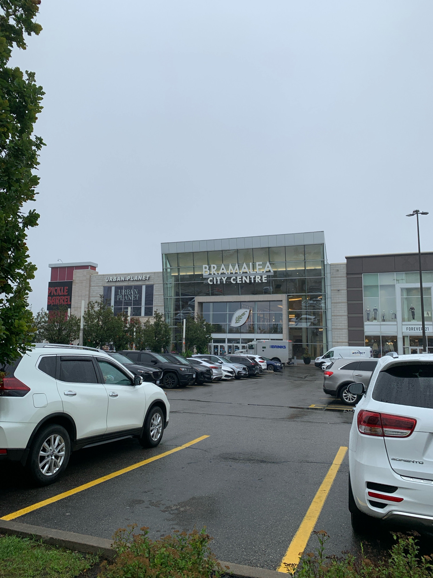 The image shows the exterior of the Bramalea City Centre in Brampton, Canada. The shopping mall features large glass windows and signage for various stores, including "Urban Planet" and "Pickle Barrel". In front of the mall, there is a parking lot filled with cars. The sky is overcast, and the ground appears wet, suggesting it may have recently rained. There are no people visible in the image. The scene is calm and depicts a typical day at a large retail complex.