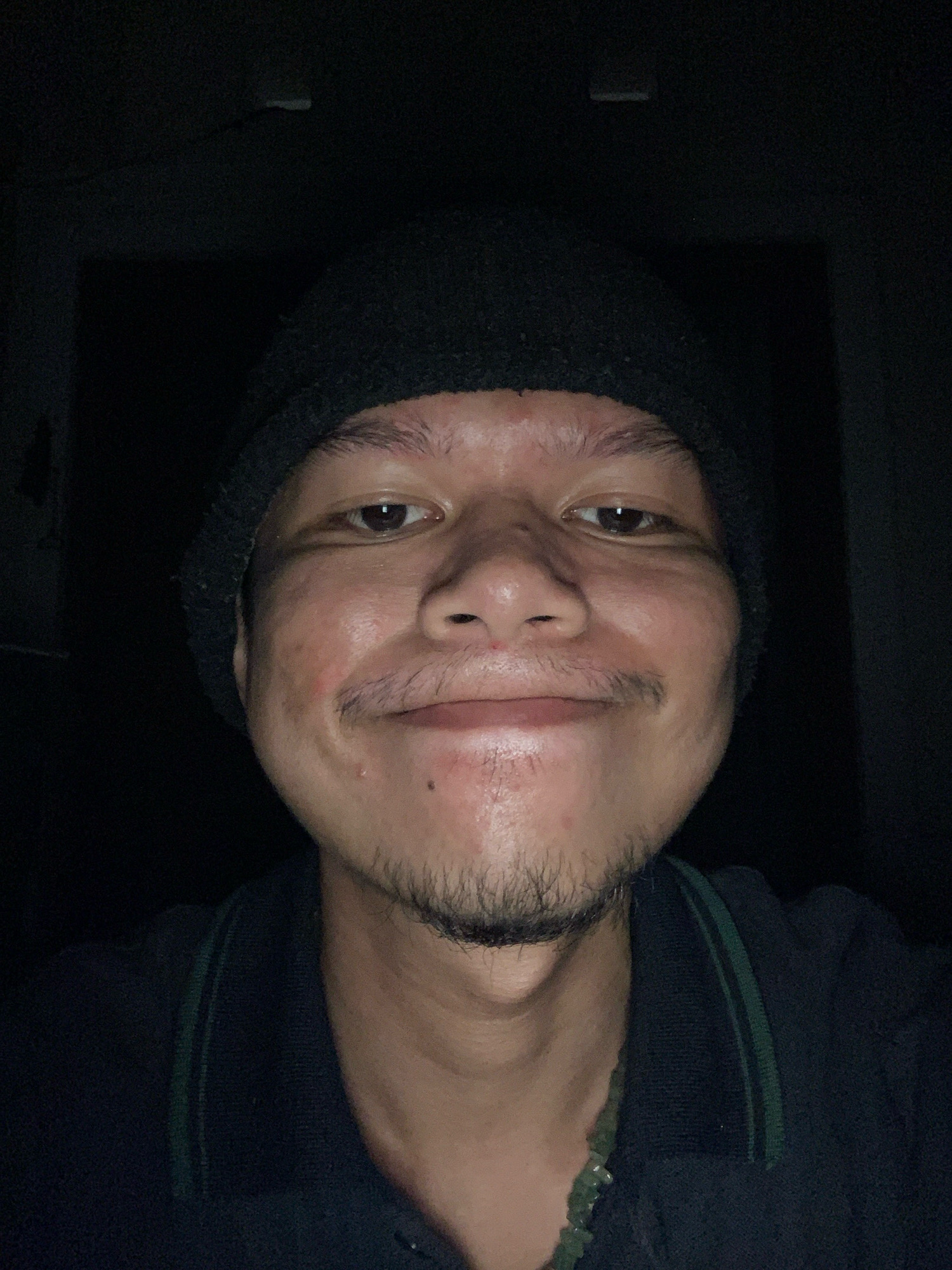 A person is captured in a close-up selfie in a dimly lit environment. The individual is wearing a dark beanie and a dark collared shirt with two thin green stripes on the collar. The person has a mustache and a goatee. The lighting suggests the photo was taken indoors, possibly at night or in a room with limited illumination. No specific landmarks of Sidoarjo, Indonesia are visible.