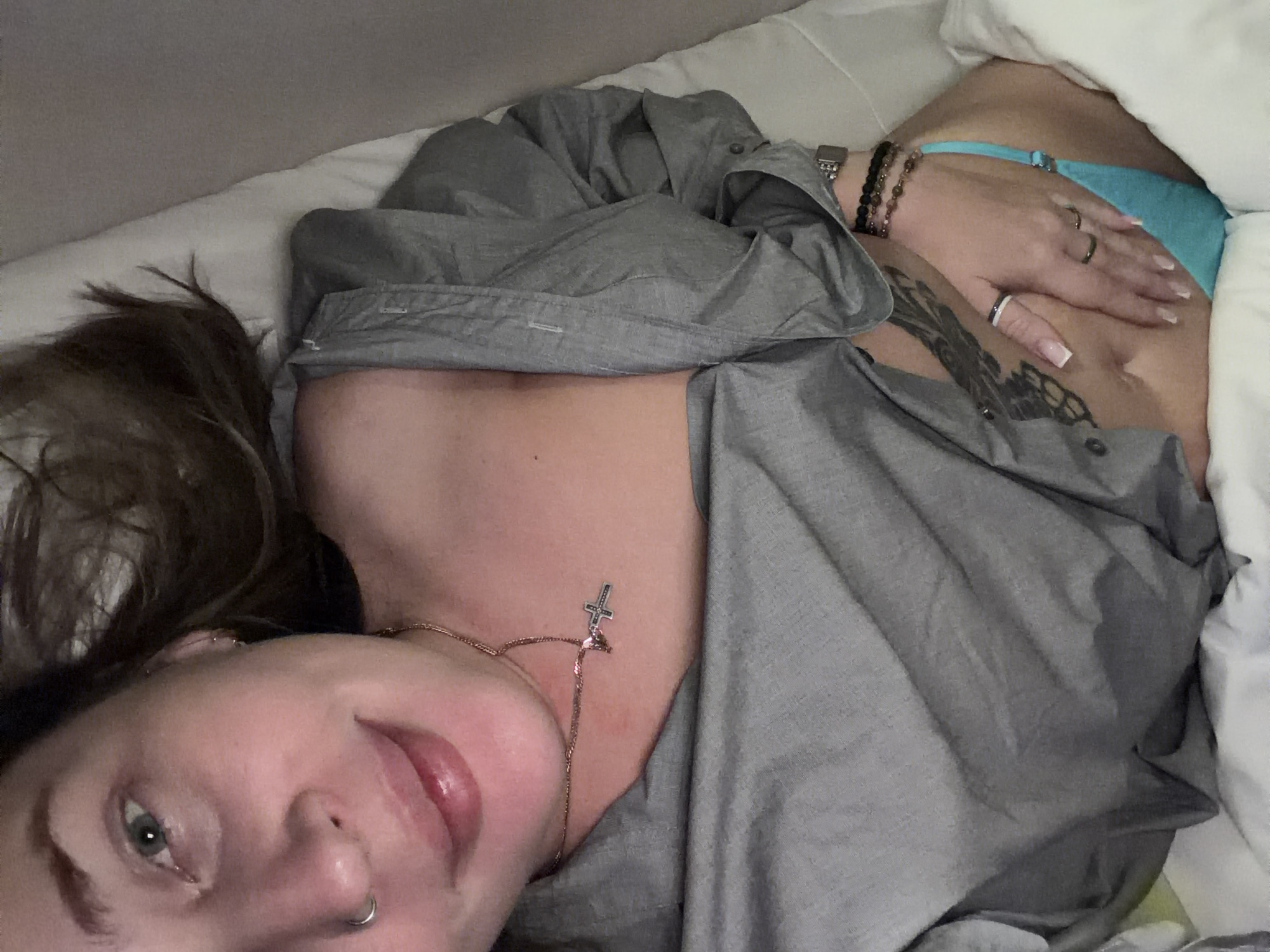 A fair-skinned person with gray-green eyes, visible facial piercings on the nose and ears, and brown hair is lying on a white bed wearing a gray button-down shirt and blue underwear. The person has tattoos on their abdomen, rings on their fingers, and bracelets on their left wrist. A gold necklace with a cross pendant is visible on the person's chest. The scene appears to be indoors, likely in a residential setting. The location in Moscow, Russia, is unknown.