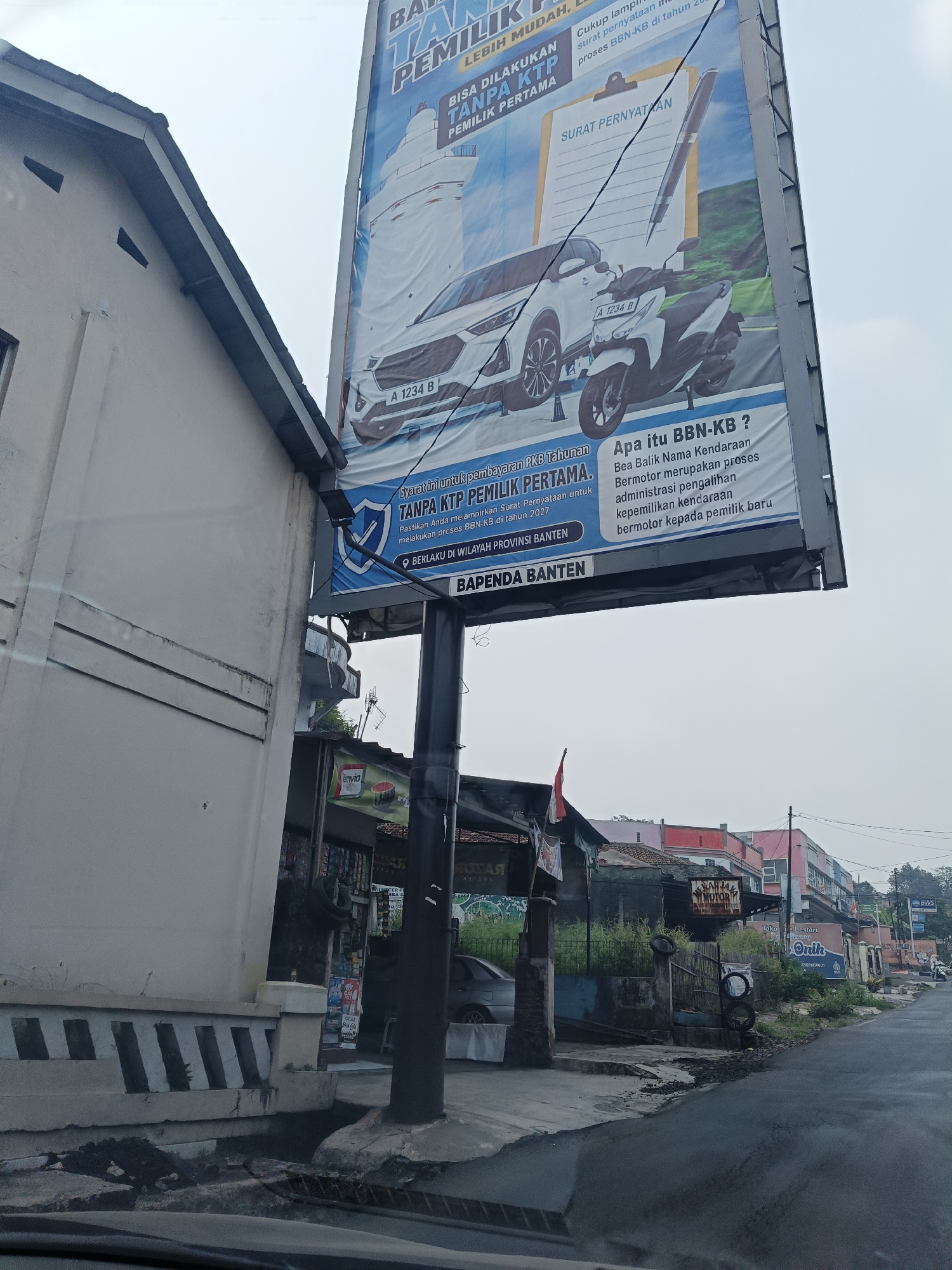 The image captures a street scene in Pandeglang, Indonesia, dominated by a large billboard advertising services related to vehicle ownership transfer ("Bea Balik Nama Kendaraan" or BBN-KB). The billboard, prominently displayed on the right side of the frame, features images of a car and a motorcycle, along with text indicating that the process can be done without a KTP (Identity Card) for first-time owners and is valid in Banten province. Below the billboard, a street with asphalt pavement runs towards the background, lined with buildings. A white building with a tiled roof is situated on the left, partially obscuring the lower portion of the billboard. In the mid-ground, various small shops and businesses are visible, with one sign reading "Banga Motor." Vegetation and some litter are present on the sidewalks and along the roadside. The sky is overcast, suggesting a cloudy day. The overall scene depicts a typical urban street with commercial activity and public service advertisements.
