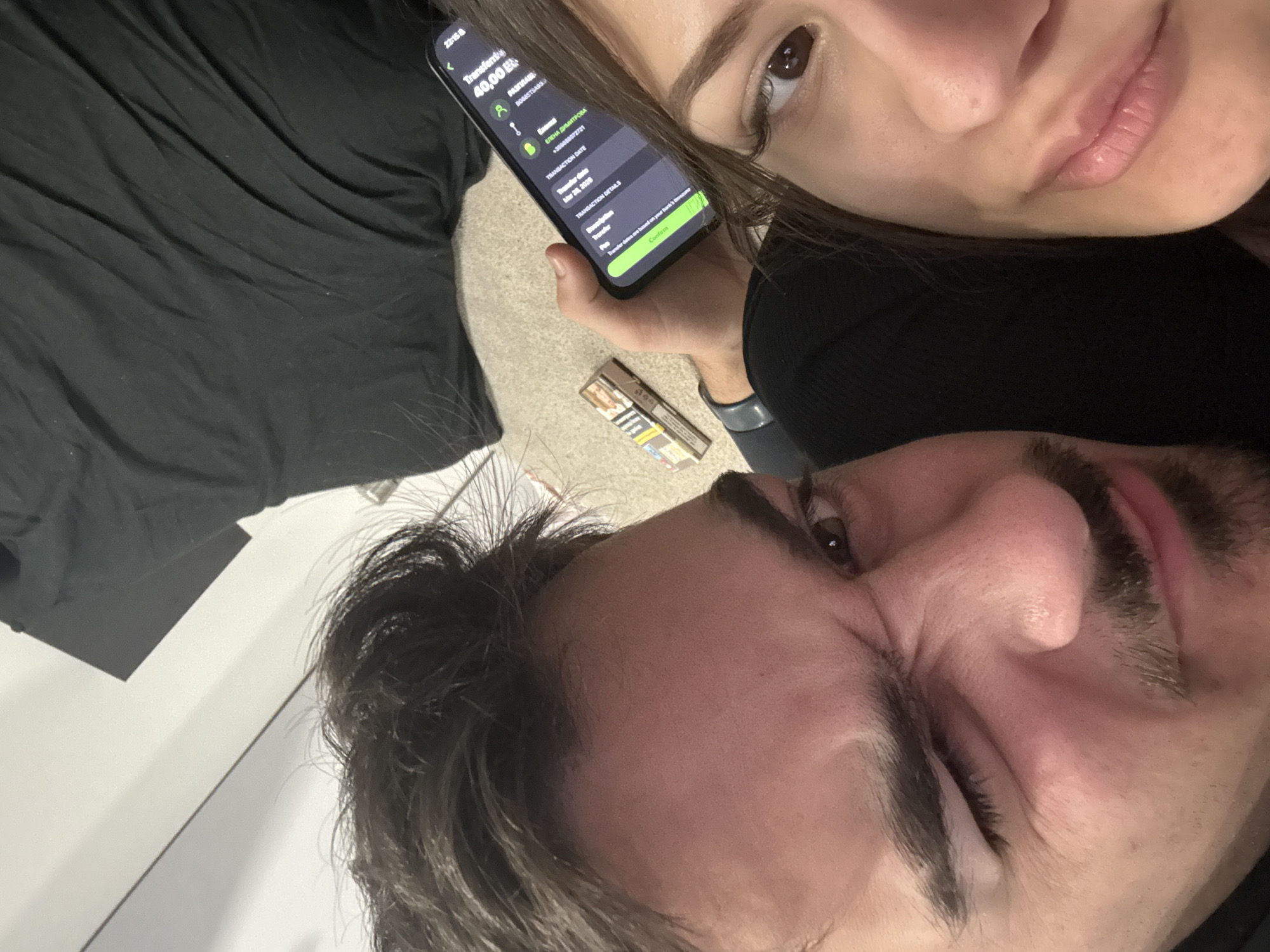 The image is a selfie taken by two individuals indoors. A man with dark hair, eyebrows, and a mustache is in the foreground. A woman with dark hair and eyebrows is positioned above him. She holds a smartphone displaying a transaction for 40.00 EUR. The recipient is listed as Elena Dimitrova with the phone number +359888072721. The transaction date is March 28, 2020. To the left of the woman is a pack of cigarettes. The background includes dark clothing and a white surface. No information is available to place the location in Sofia, Bulgaria.