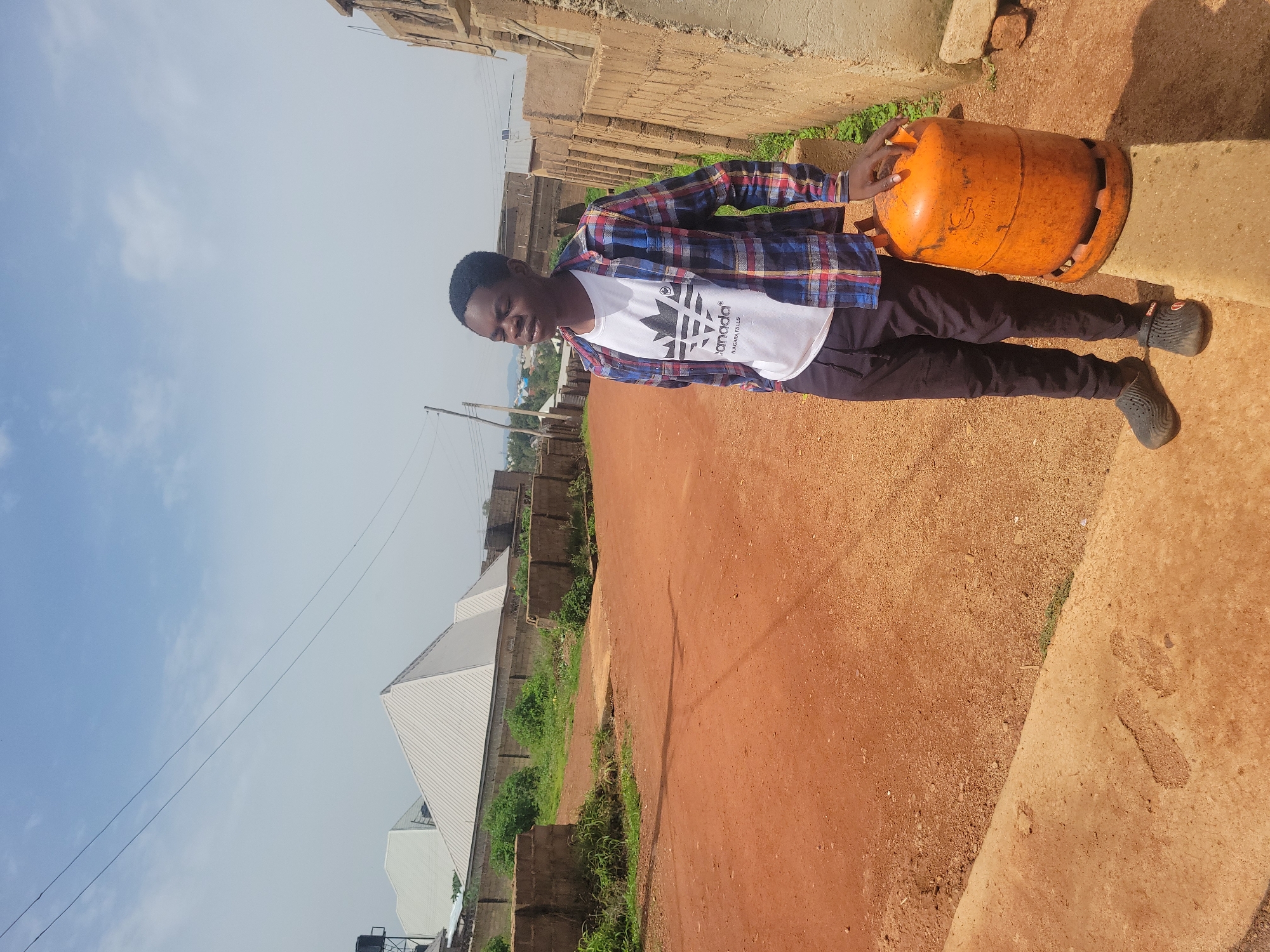 A young Black male is pictured standing outdoors, in what appears to be a semi-urban area. The background suggests the city of Jos, Nigeria, with its characteristic red earth and brick structures. He is wearing a blue, red, and white plaid shirt over a white t-shirt with a black graphic print. He is also wearing dark-colored pants and dark slip-on shoes. The male is positioned to the right of the frame and is holding an orange propane gas cylinder with his right hand. The gas cylinder is lying on its side on the ground. The ground is covered in reddish-brown earth, with some patches of green grass and low-lying bushes visible. In the background, there are unfinished brick buildings and a cloudy sky. The orientation of the image is such that the male is facing towards the viewer and the gas cylinder is in front of him.