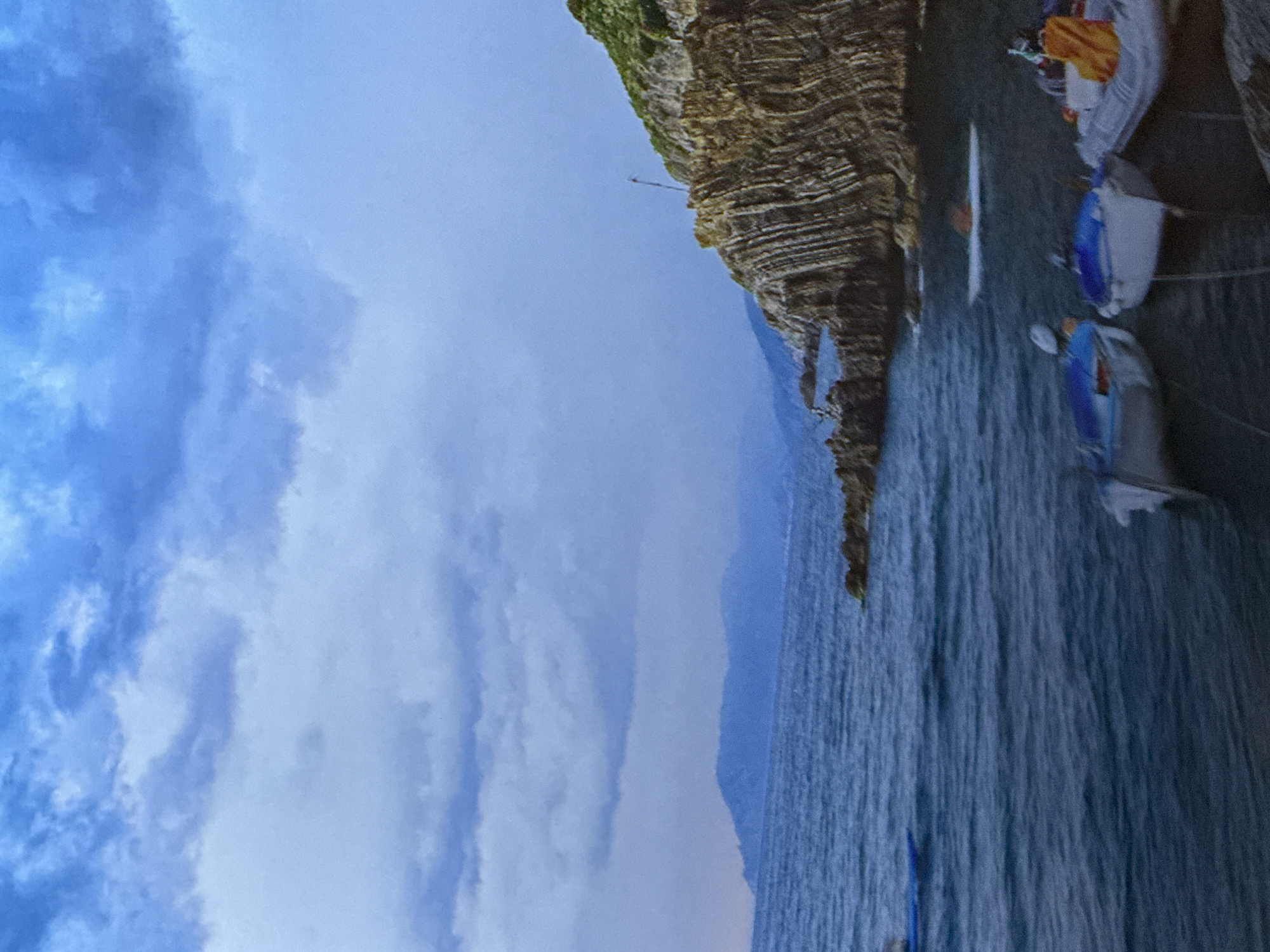 The image depicts a rocky cliff face descending into a body of water. Several small boats, painted in shades of white and blue, are moored near the cliff. Some of the boats have brightly colored items, possibly clothing or sails, draped over them. The water is a deep blue-gray, with ripples suggesting a gentle movement. Above the water, the sky is a pale blue, filled with wispy, white clouds that are beginning to gather. The rocky cliff is layered and appears to be made of sedimentary rock, with patches of green vegetation clinging to its upper edges. In the distance, hazy, bluish mountains can be seen. There is a thin, vertical structure with a red flag on top jutting out from the cliff face, possibly a marker or a small observation post. The overall scene suggests a calm, coastal environment, likely in the late afternoon or early evening, given the soft light and gathering clouds. There are no visible people. No text is present in the image.