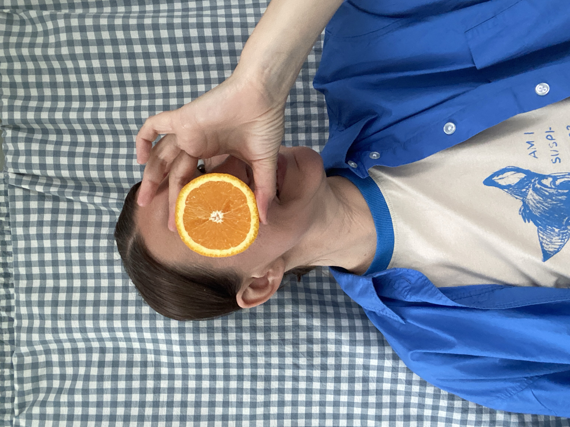 A person lies on a blue and white checkered surface. They are wearing a blue shirt over a white t-shirt with a blue design. An orange slice is held up in front of their eye. The location is unspecified and there is no indication of it being in Chaikovsky, Russia.