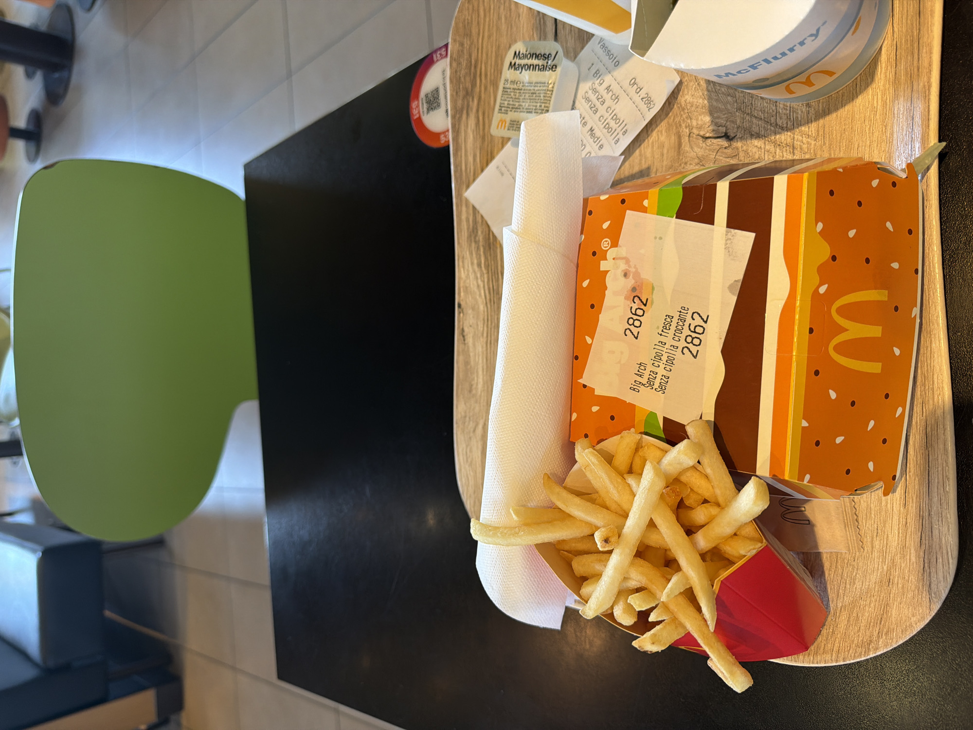 The image depicts a McDonald's meal on a table. The location is likely in Capiago Intimiano, Italy, based on the language on the packaging ("Senza cipolla," "Maionese/Mayonnaise"). The meal consists of a Big Mac in its packaging, a container of french fries, a McFlurry, napkins, a mayonnaise packet, and receipts. The fries appear to be partially eaten. The order number "2862" is prominent. A green chair is visible in the background. The table is black and the tray is light wood-colored.