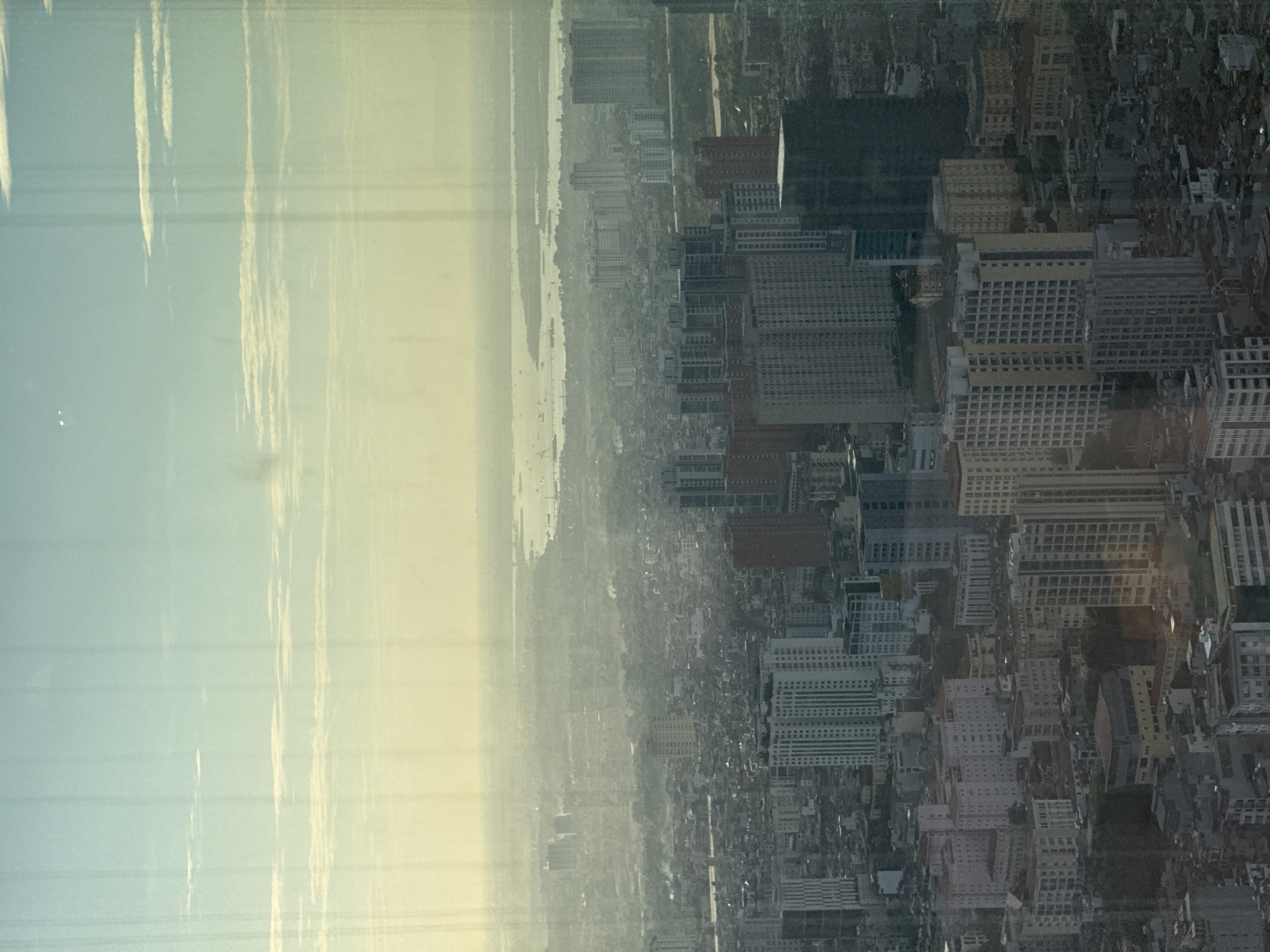 This is a high-angle, wide shot of a dense urban landscape in Hanoi, Vietnam, seen through a window with vertical smudges. The sky above is a pale blue and yellow gradient, with thin, wispy clouds. The horizon is hazy, with distant bodies of water dotted with boats and a faint landmass. The mid-ground of the image showcases a sprawling city with numerous buildings of varying heights and architectural styles, some appearing modern high-rises and others older, lower-rise structures. There is a notable presence of tall apartment buildings, many with grid-like window patterns. The foreground is filled with more densely packed buildings, creating a sense of an expansive and busy metropolitan area. The overall impression is one of urban sprawl under a hazy sky, likely during daylight hours given the sky's coloration and the illumination of the buildings.