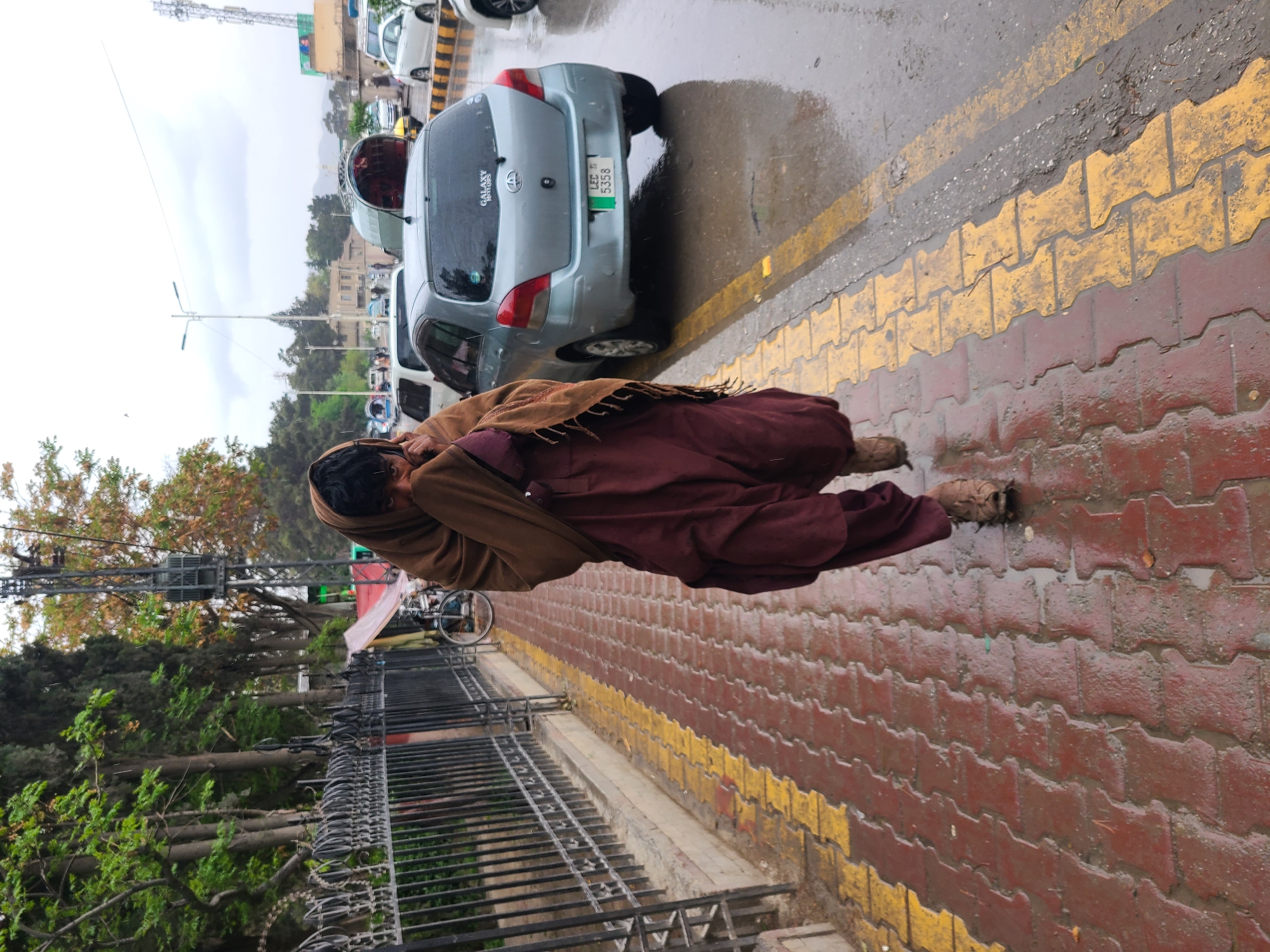 The image depicts a pedestrian in Quetta City Tehsil, Pakistan, walking on a rain-soaked brick sidewalk next to a road. The person is wrapped in a brown shawl and wears maroon clothing. Cars are parked along the side of the road. A black metal fence separates the sidewalk from a green area with trees and possibly a park. Power lines and a communications tower are visible in the background. The weather appears overcast and rainy.