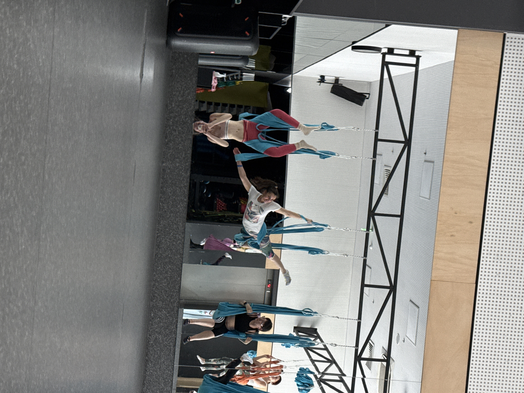 The image shows a group of individuals participating in aerial yoga, suspended from blue fabric hammocks. The location appears to be an indoor gym or studio with mirrored walls reflecting the scene. The participants are of varying ages and attire, some in athletic wear and others in more casual clothing. They are engaging in various poses and stretches within the fabric. The background includes gym equipment, flooring, and structural support for the aerial apparatus.
