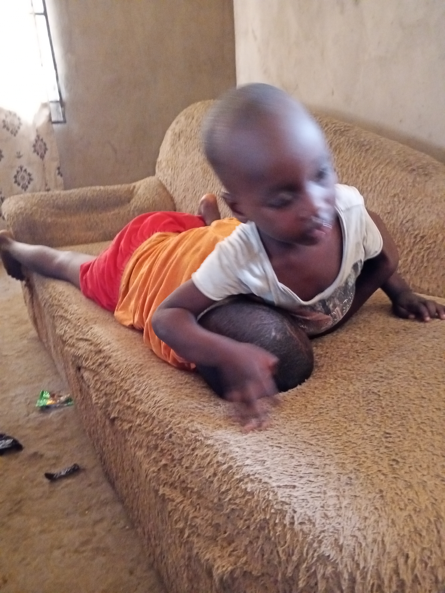 The image depicts two young children indoors, engaged in play or rest on a large, light brown, textured sofa. In the foreground, one child, wearing a white t-shirt, is positioned on all fours or kneeling on the couch, with their head blurred by motion and facing towards the bottom left. The second child is lying prone on the sofa, partially beneath or beside the first, dressed in red shorts and an orange top, with their legs stretched out. Their head is also motion-blurred and partially visible.
The setting appears to be a modest indoor space, likely a home. Bright daylight streams in through a window visible on the left side of the frame, suggesting it is daytime. The walls are a light brown or beige color, and there's a subtle patterned fabric or wallpaper in the background near the window. On the floor next to the couch, several small pieces of litter, including a green wrapper and a dark crumpled item, are visible. The sofa itself has a rough, somewhat shaggy texture and shows signs of use. There is no visible text in the image.