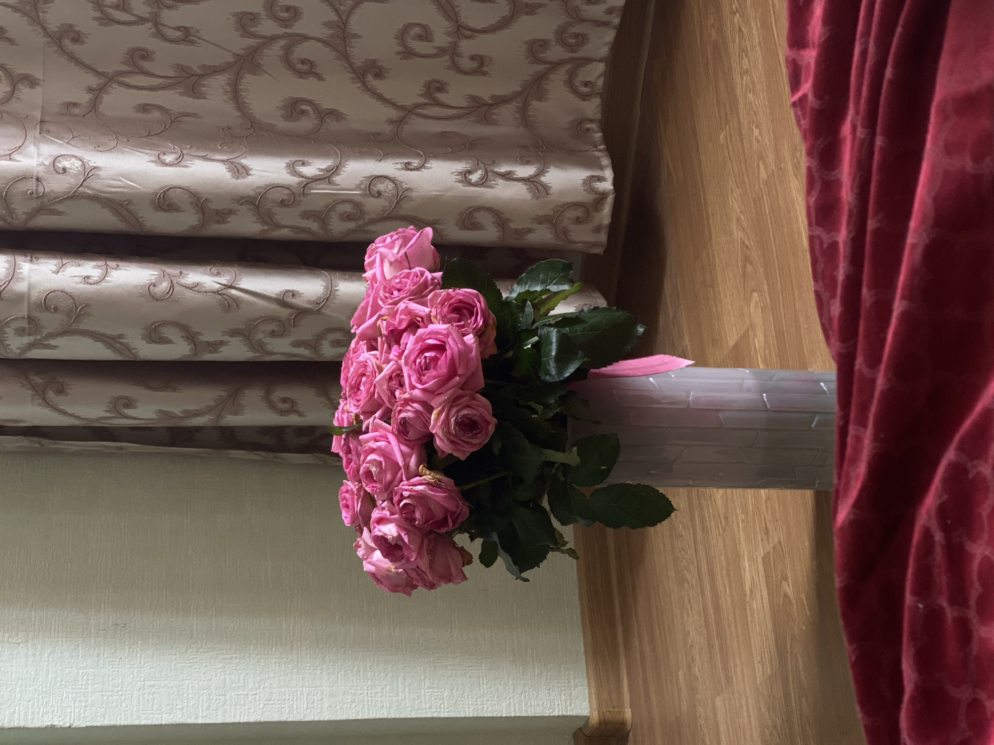 A bouquet of pink roses is arranged in a container, sitting on a shelf or surface in front of a window covered by a patterned blind. A dark red curtain is draped to the right of the roses. The interior setting includes wood-patterned wall panels and a textured wall. The location, Sochi, Russia, is assumed and not directly evident from the image. There are no people visible in the image.
