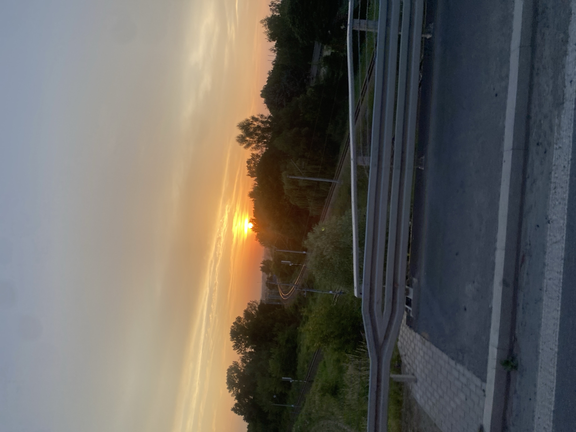 This media file depicts a sunset scene with a railway line winding through a wooded area. The sun is low on the horizon, casting a warm, orange glow across the sky and illuminating the clouds with golden hues. The trees are dense and green, suggesting a natural, possibly rural, setting. A metal railing and a paved surface, possibly a walkway or roadside, are visible in the foreground, indicating that the vantage point is from a raised structure overlooking the landscape, such as a bridge or overpass. The railway tracks are clearly visible, curving into the distance, with overhead electrical wires and posts indicating it's an active line. The overall mood is serene and peaceful, evoking the beauty of nature at the end of the day. The time of day is clearly sunset, and the weather appears clear with some cloud formations catching the light. There is no visible text in the image.