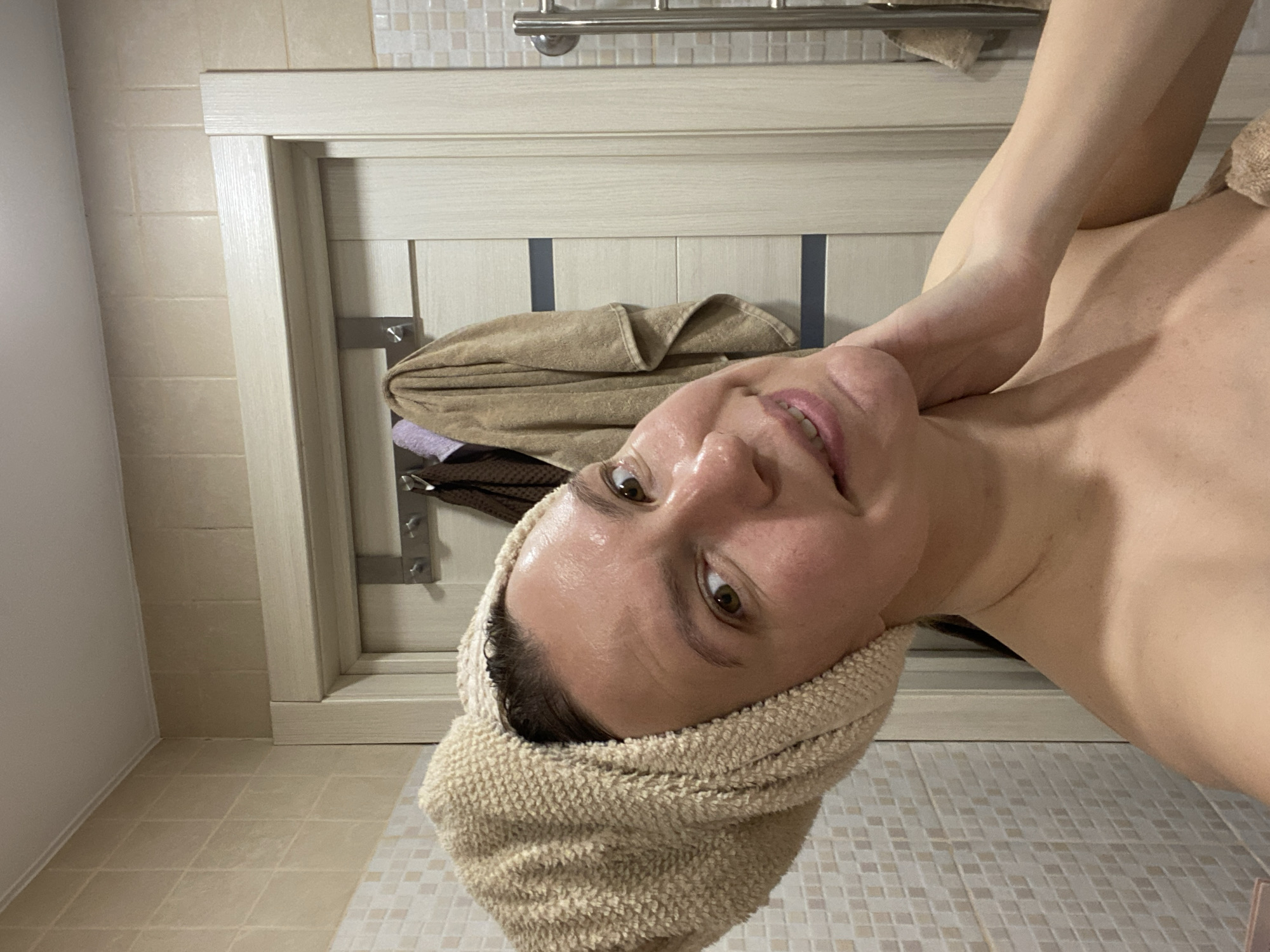 A woman with a towel wrapped around her head stands in a bathroom. The bathroom has beige and white tiled walls and floors. A towel rack with towels hangs on the wall behind her. The location is not discernible as Chaikovsky, Russia.
