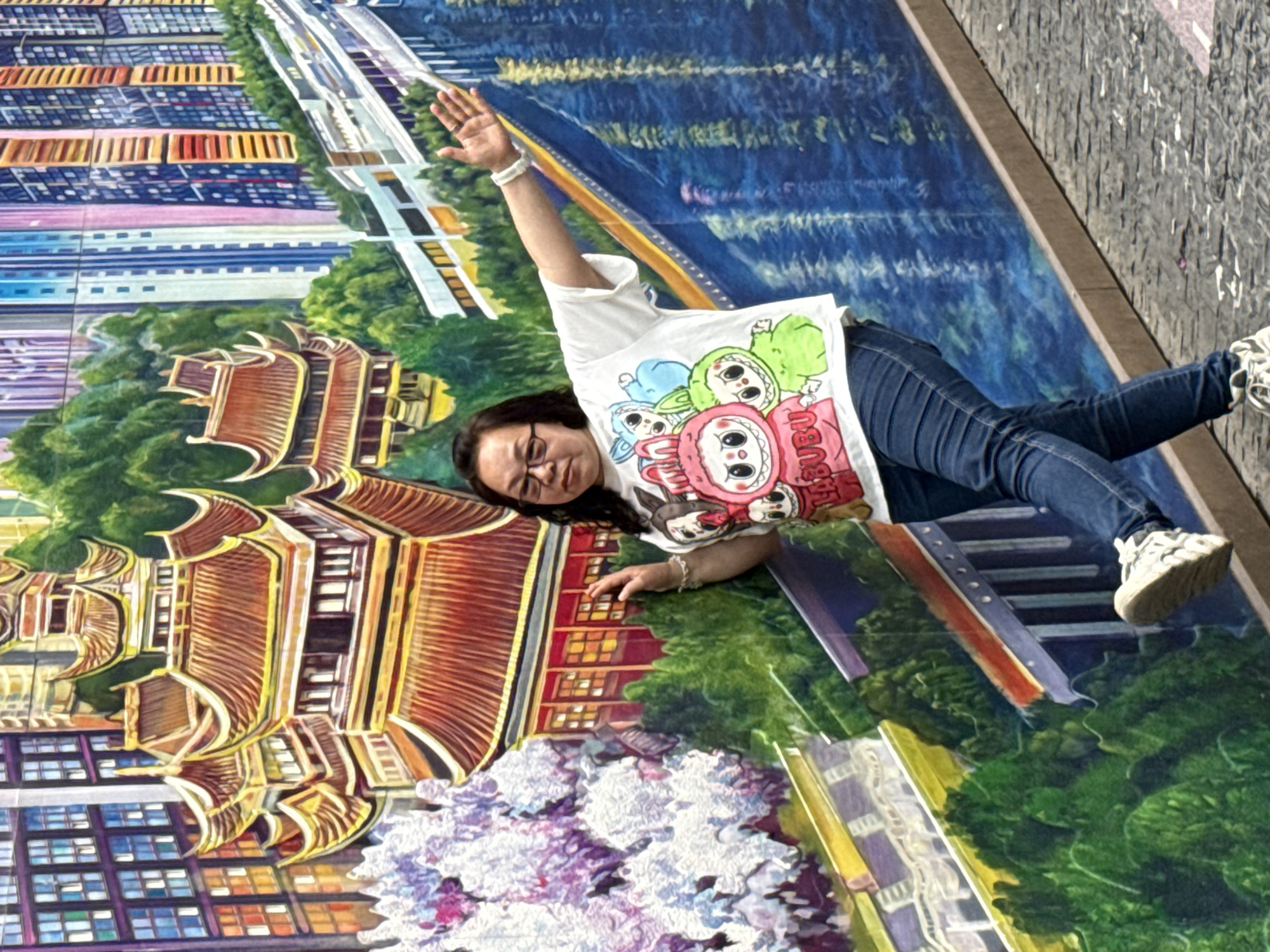 A woman wearing glasses, a white shirt with cartoon characters, and blue jeans is lying on a street mural in Yuzhong District, China. She has one arm raised. The mural features an aerial view of a city, including buildings, trees, a river, and a bridge. The background shows a gray asphalt area.