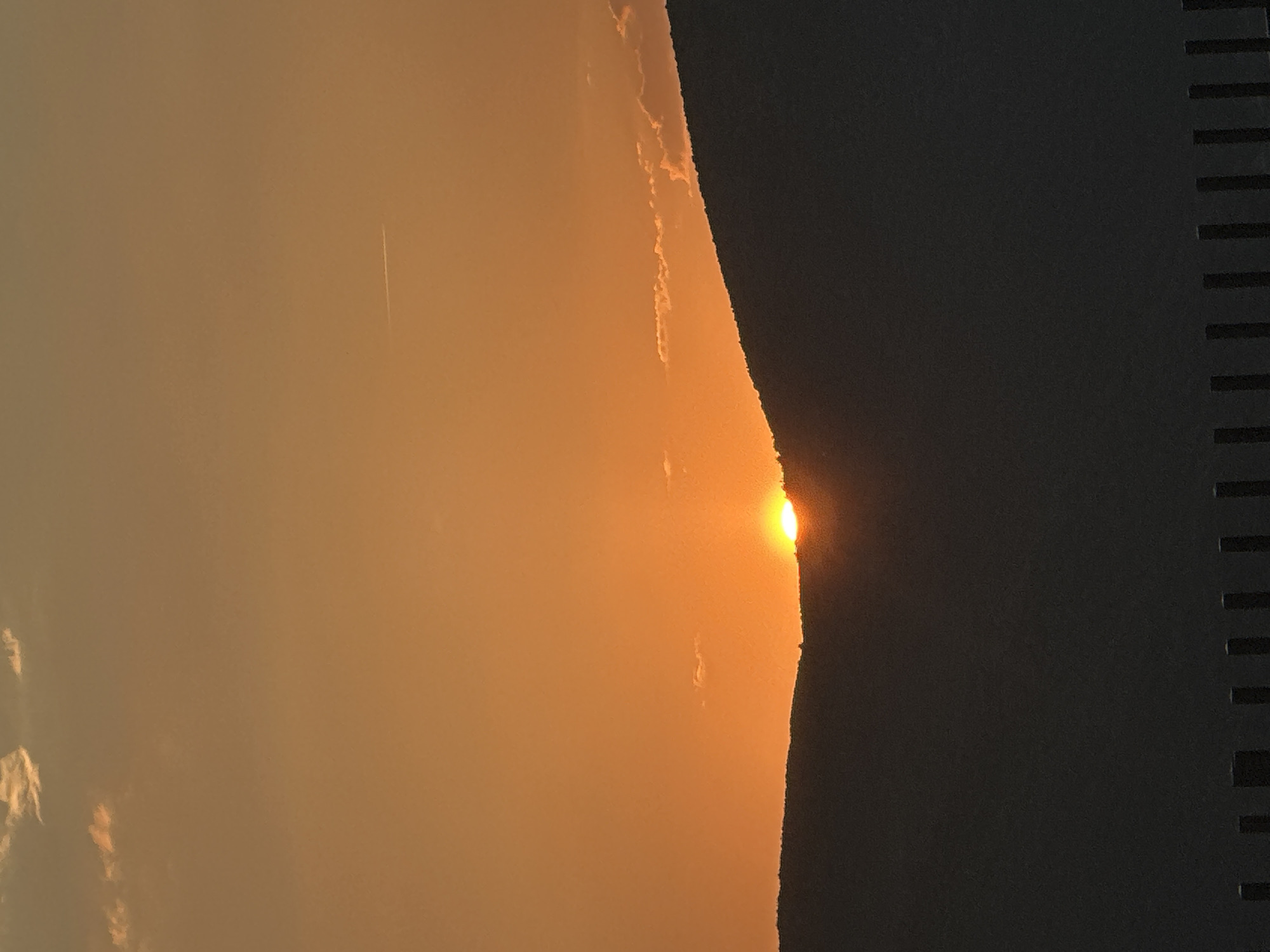 The image shows a sunset over what appears to be a building or structure, possibly in Budva, Montenegro, judging from the location. The left side of the image displays a hazy orange sky, with some indistinct cloud formations. The sun is partially obscured by a dark object, likely a building, with a bright glow at the point of contact. To the right of the glowing sun is a dark, shadowy section with regularly spaced horizontal lines, suggesting a part of a building's facade or window structure. A vapor trail of an airplane is seen in the orange sky.