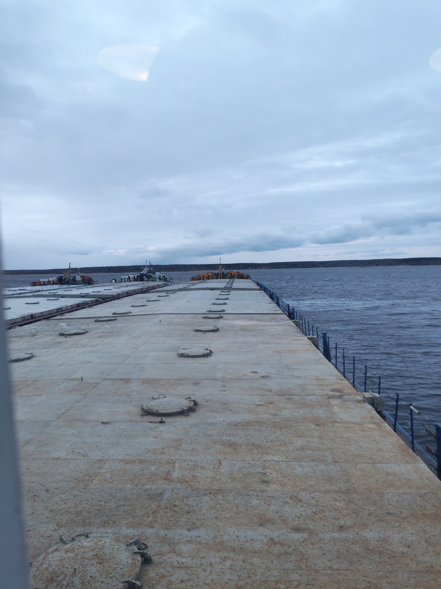 The image captures a wide-angle view of a large, flat, concrete or metal structure, possibly a pier, barge, or floating platform, extending into a body of water under an overcast sky. The structure dominates the foreground and midground, running from the bottom left to the upper right of the frame, tapering slightly in the distance. Its surface is light gray and appears weathered, with numerous round, metal hatch covers evenly spaced across it. Along the right edge of this structure, a blue metal railing or fence runs parallel to the water's edge.
To the left of the main structure, further out in the water, several smaller vessels resembling tugboats or workboats are visible. These boats are low-lying, with superstructures that are light in color, possibly white or light gray, with some yellow and green accents.
The body of water, likely a river or large lake, appears dark blue-gray with visible ripples, suggesting a light breeze. A distant landmass, covered with trees, forms the horizon line. The sky is heavily overcast with varying shades of gray clouds, indicating a cloudy day. There are no people visible in the image. A bright, undefined light reflection or glare is present in the upper left portion of the sky. No discernible text is visible. The scene conveys a utilitarian or industrial waterside setting.