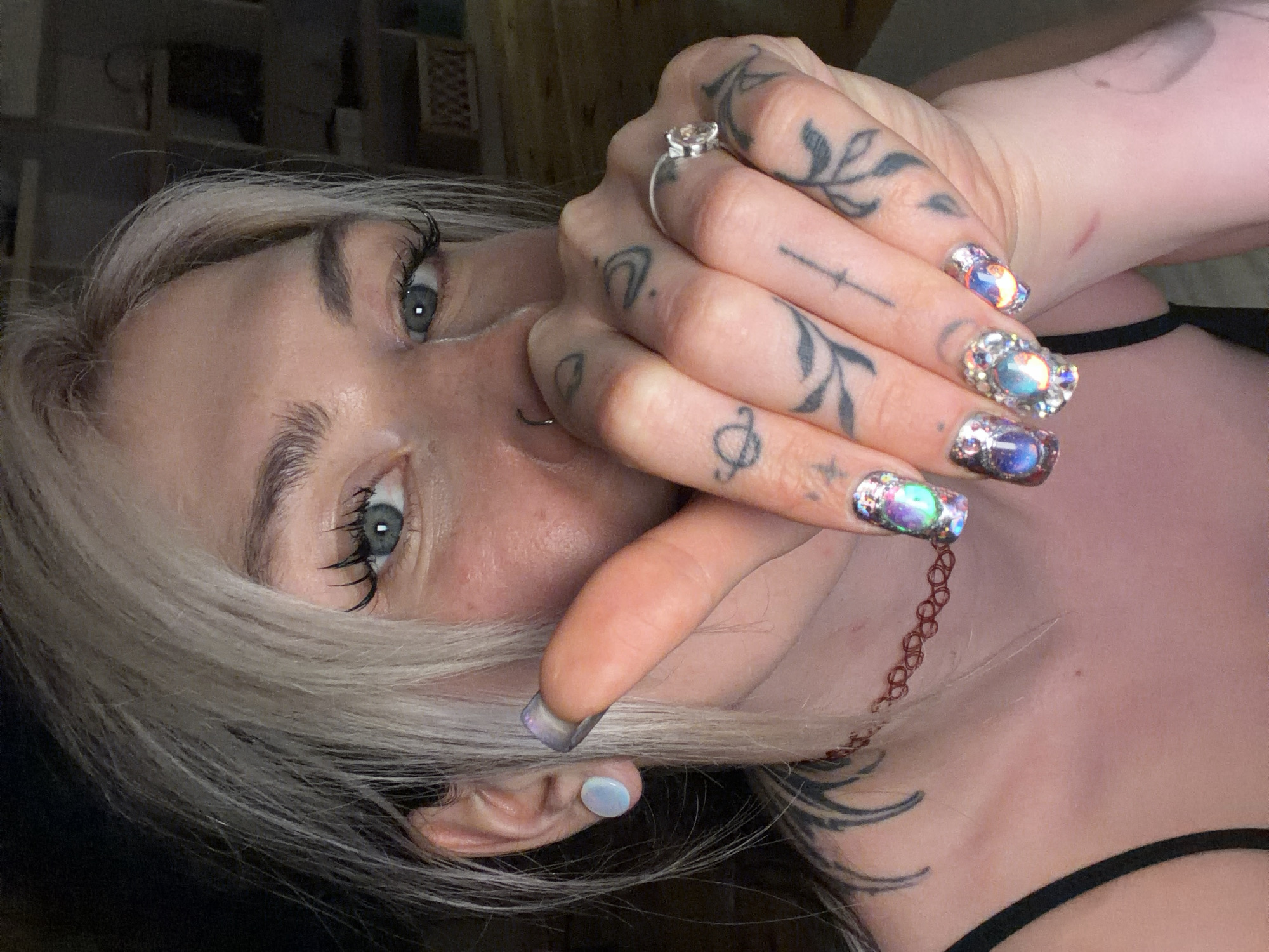 A fair-skinned person with platinum blonde hair is pictured close-up. They have light blue eyes with dark eyelashes. Their left hand is raised near their face, displaying several tattoos on their fingers and hand, including a ring on their ring finger. The person is wearing a black top, and a red choker necklace is visible on their neck. The background is dark and somewhat indistinct. The location is not provided.