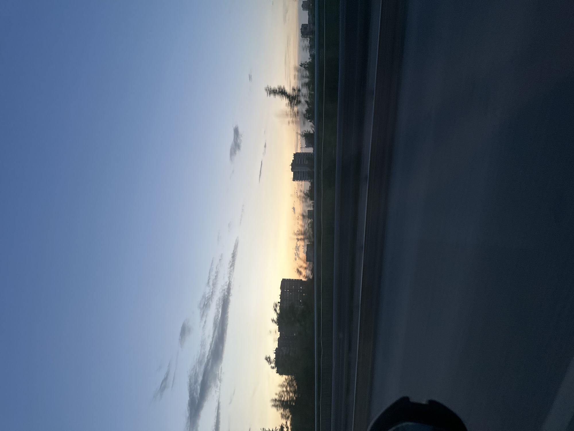 The image depicts a view from a vehicle on a highway or elevated roadway, likely at dusk or dawn. To the left, the sky is visible, transitioning from a deeper blue to a lighter, almost orange hue near the horizon. Several elongated clouds stretch across the sky. In the distance, the silhouettes of buildings can be seen, partially obscured by trees. A metal guardrail runs along the right side of the road, and the surface of the roadway is visible, blurring due to motion. The lower part of the frame contains part of the interior of the vehicle, possibly the dashboard and a rear-view mirror. It is not possible to confirm if the location of this photo is in the city of Perm, Russia with the information provided.