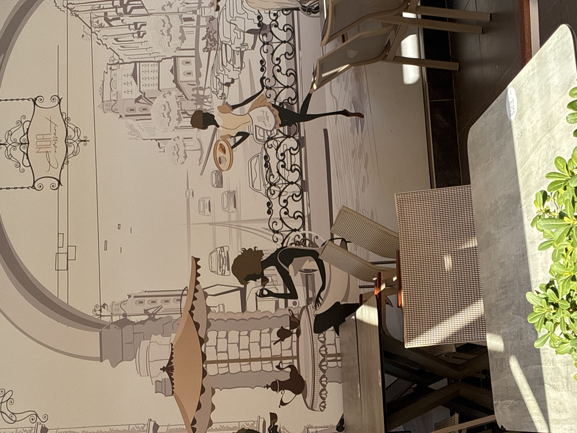 The image depicts a mural on a wall in a cafe, likely in Alanya, Turkey. The mural features a stylized black and white scene of a European street with a Parisian cafe atmosphere. In the foreground of the mural, a silhouette of a woman sits at a table under an umbrella, drinking from a cup. Further back, another silhouette of a woman, possibly a waitress, walks by carrying a tray with a croissant and a coffee. The background of the mural shows buildings, parked cars, and a decorative fence. In front of the mural, real-world elements of the cafe are visible, including tables and chairs. Sunlight casts shadows on the tables and chairs in the foreground. The mural itself has a sign at the top that reads "NOA Croissant." The cafe appears to be an outdoor or semi-outdoor seating area, given the presence of tables and chairs in the sunlight and the mural's design suggesting an outdoor cafe. The overall impression is of a charming and artistic cafe environment.
