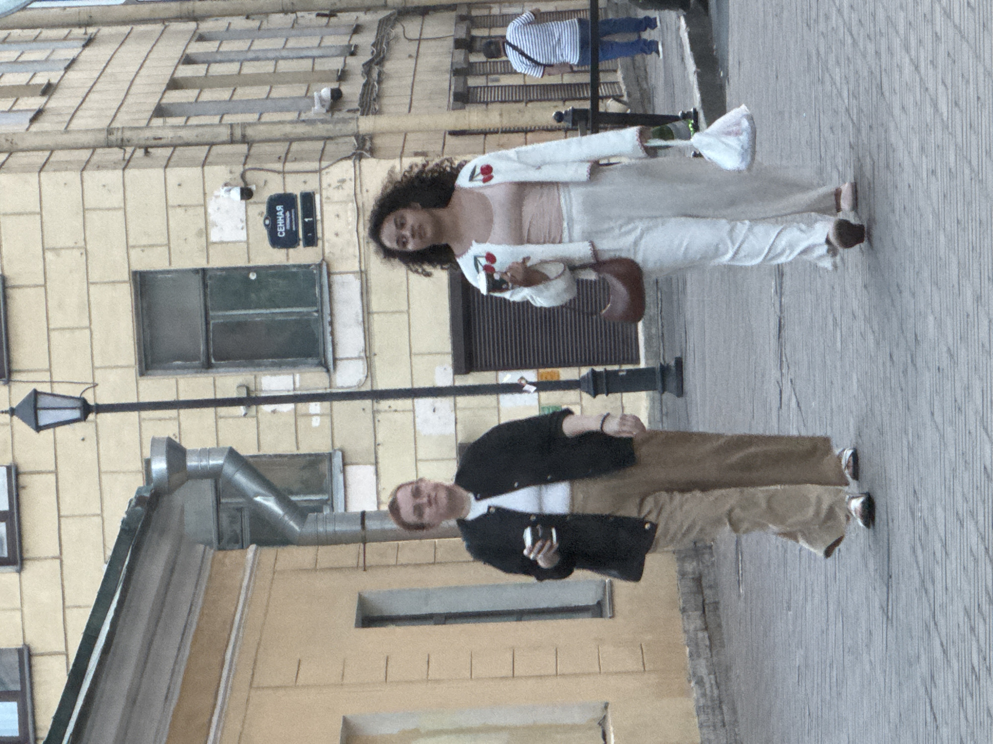 This image depicts a daytime street scene in an urban area, identified as Saint Petersburg, Russia, given the provided context. The ground is paved with light-colored blocks, and buildings with multiple windows line the street.
In the foreground, two individuals are prominent. On the left (after rotating the image), a person with reddish hair wears a black jacket over a white shirt and tan trousers, holding a smartphone in their right hand. On the right, a person with dark, curly hair is dressed in a white cardigan embroidered with red cherries, a light pink top, and white pants. This individual holds a brown, saddle-shaped bag or purse and what appears to be a phone. Both seem to be standing casually on the sidewalk.
Further back, a third person in a striped shirt and blue pants is walking away. The surrounding buildings are light-colored, featuring various windows, utility lines, and ventilation pipes. A black lamp post is also visible.
A notable detail is a dark blue street sign mounted on a building, which clearly displays the text "СЕННАЯ" (Sennaya) at the top, "ПЕРЕУЛОК" (Pereulok, meaning lane or alley) below it, and the number "1". This identifies the location as Sennaya Pereulok 1. The lighting is soft and even, suggesting it might be an overcast day.