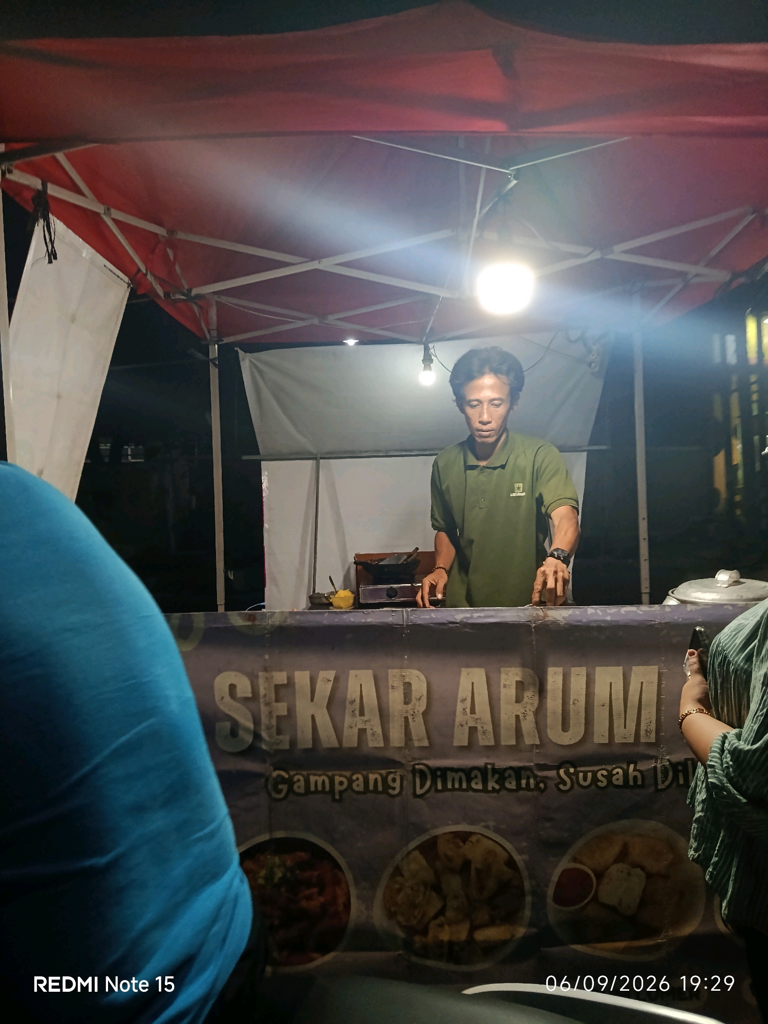 A street food vendor in a green polo shirt stands behind a food stall at night in Keboansikep, Indonesia. The vendor appears to be male, with dark hair and a focused expression, and is reaching out towards the counter. He is wearing a wristwatch on his left wrist. The stall has a red and white canopy overhead, illuminated by a bright light. A banner with the text "SEKAR ARUM Gampang Dimakan, Susah Dil" is displayed on the front of the stall, with images of food below. The environment is dark, suggesting it is nighttime. A person in a blue shirt is partially visible in the foreground on the left, and another person wearing a green striped garment is on the right. The bottom of the image shows a timestamp "06/09/2026 19:29" and "REDMI Note 15".