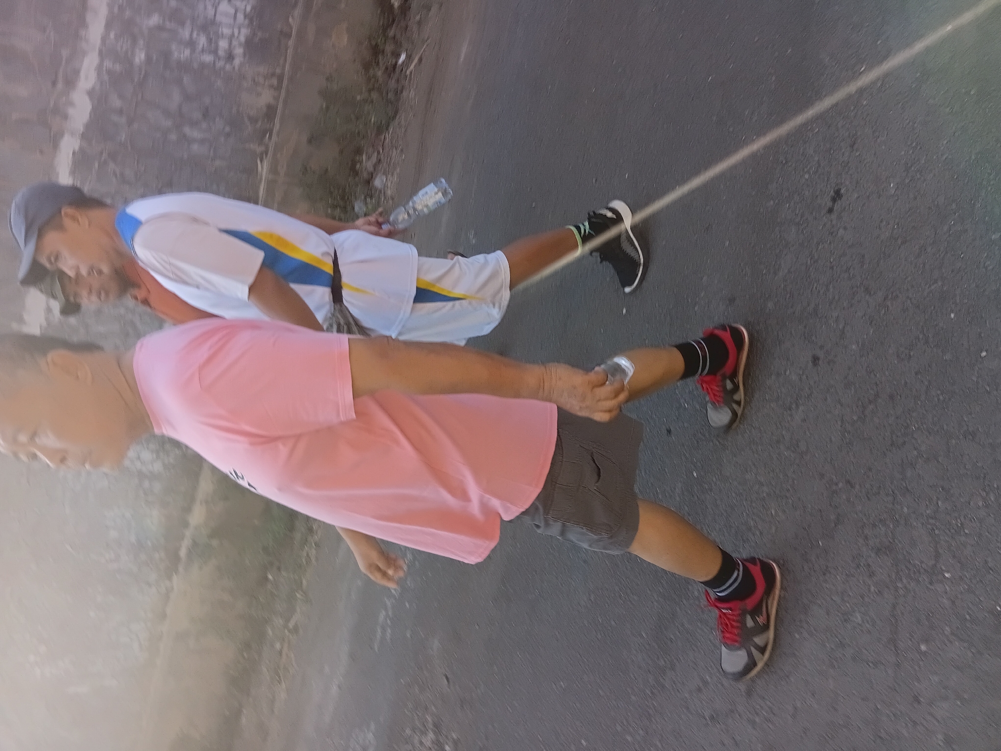 Three individuals are walking on an asphalt road in Tanggul Wetan, Indonesia. The person in the foreground, on the left, is wearing a pink t-shirt and grey shorts with red and black athletic shoes. The person in the middle is wearing a white shirt with blue and yellow accents and white shorts with black athletic shoes. The person in the background is wearing an orange shirt and a hat. The environment appears to be outdoors, with a textured wall on the right side of the image. The lighting suggests it is daytime.
