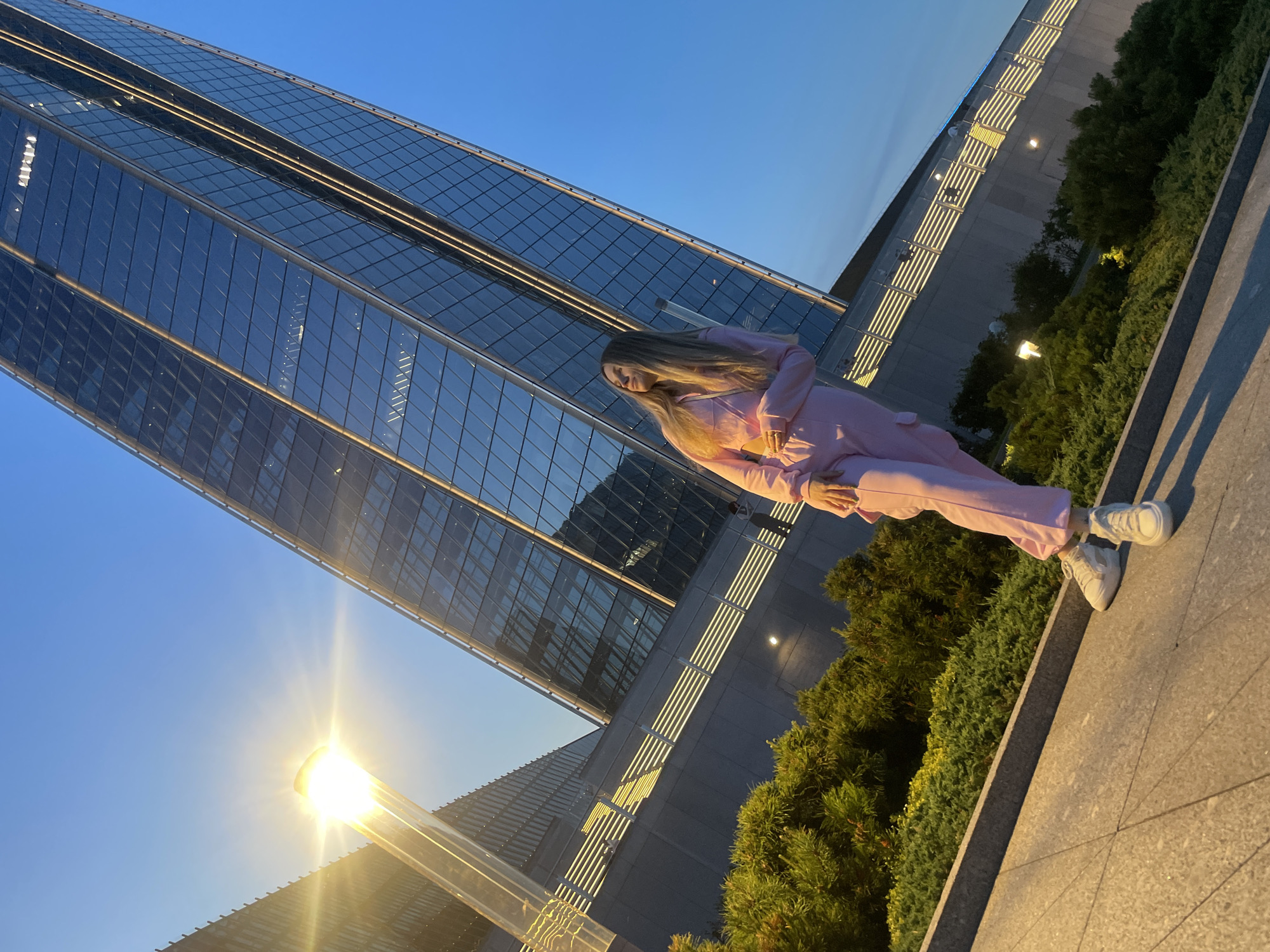 An outdoor, full shot captures a young woman with long blonde hair standing on a paved surface in front of a modern, glass-facade skyscraper. The woman is dressed in a pink tracksuit and white sneakers, her body angled towards the right of the frame as she gazes upward. Her left hand is raised, resting near her face. The skyscraper, a dominant element of the background, reflects the bright sky and some of the surrounding greenery. The sky is a clear, vibrant blue, suggesting either early morning or late afternoon. To the left of the frame, a tall, modern lamppost emits a bright, warm light, casting a strong glow and creating a lens flare effect. In the foreground, lush green bushes and manicured shrubbery line a grey stone border. The overall scene suggests an urban setting, possibly a public plaza or park adjacent to an office building, in Saint Petersburg, Russia.