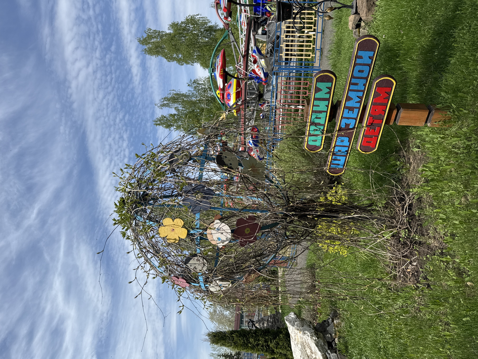 The image shows an outdoor scene in Chaikovsky, Russia. A sign with the words "Для Земли Детям" (For the Children of the Earth) is visible in the foreground. Behind the sign, there's a small tree or shrub decorated with cut-out flowers. A carousel is visible in the background. The ground is covered in green grass. The sky is blue with streaky clouds.