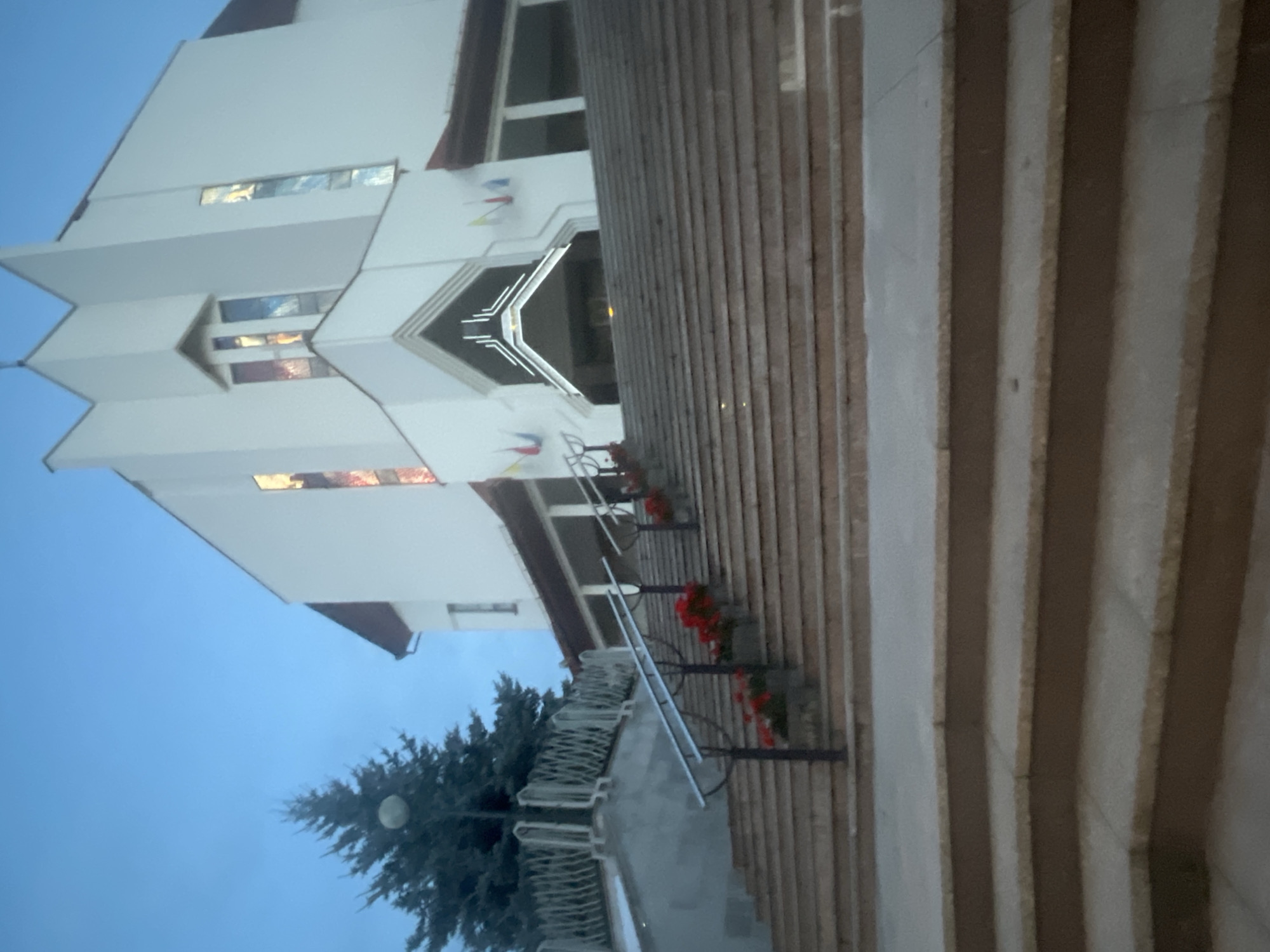 The image shows the exterior of a modern church building with a tall, white facade and a prominent set of stairs leading up to the entrance. The sky is a pale blue, suggesting it is either early morning or late evening. To the left of the church, there is a tall evergreen tree. Along the sides of the stairs, planters with red flowers add a touch of color. The architectural style of the church is contemporary, with clean lines and angular elements, including stained glass windows visible on the upper portion of the facade. The scene appears to be devoid of people and any significant activity, presenting a quiet and serene atmosphere. This building is identified as the Church of the Divine Mercy in Białystok, Poland.