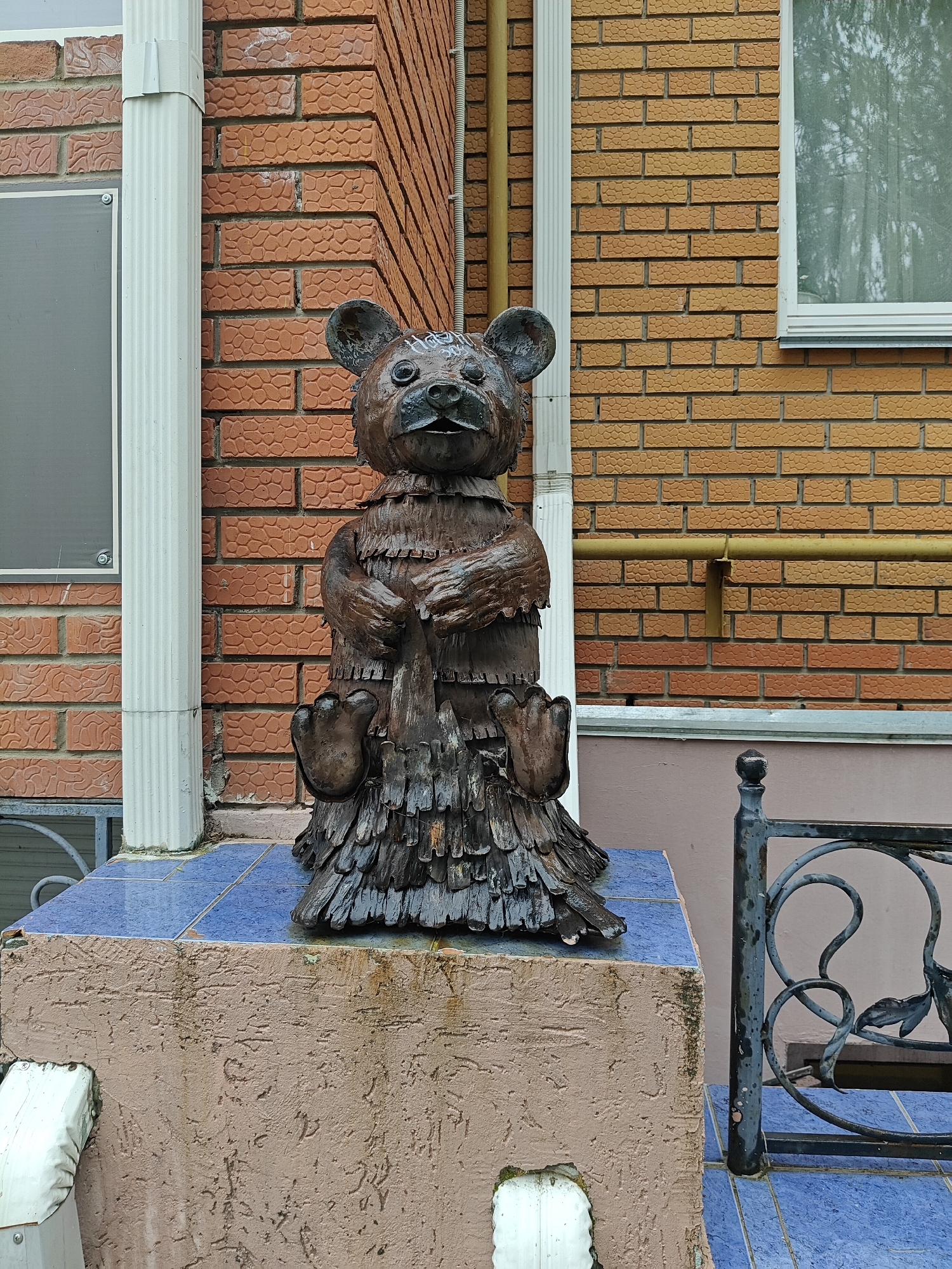 A bronze statue of a bear sits atop a tiled pedestal in front of a brick building. The bear is stylized with rough, layered features, resembling wood carvings. It sits upright, clasping a long object to its chest, perhaps a tool or a fish. The building has both red and tan brick sections, with downspouts running along the corners. The pedestal is tiled in blue and sits on a textured base. A decorative metal railing is partially visible to the right of the statue. The location is Chaikovsky, Russia.