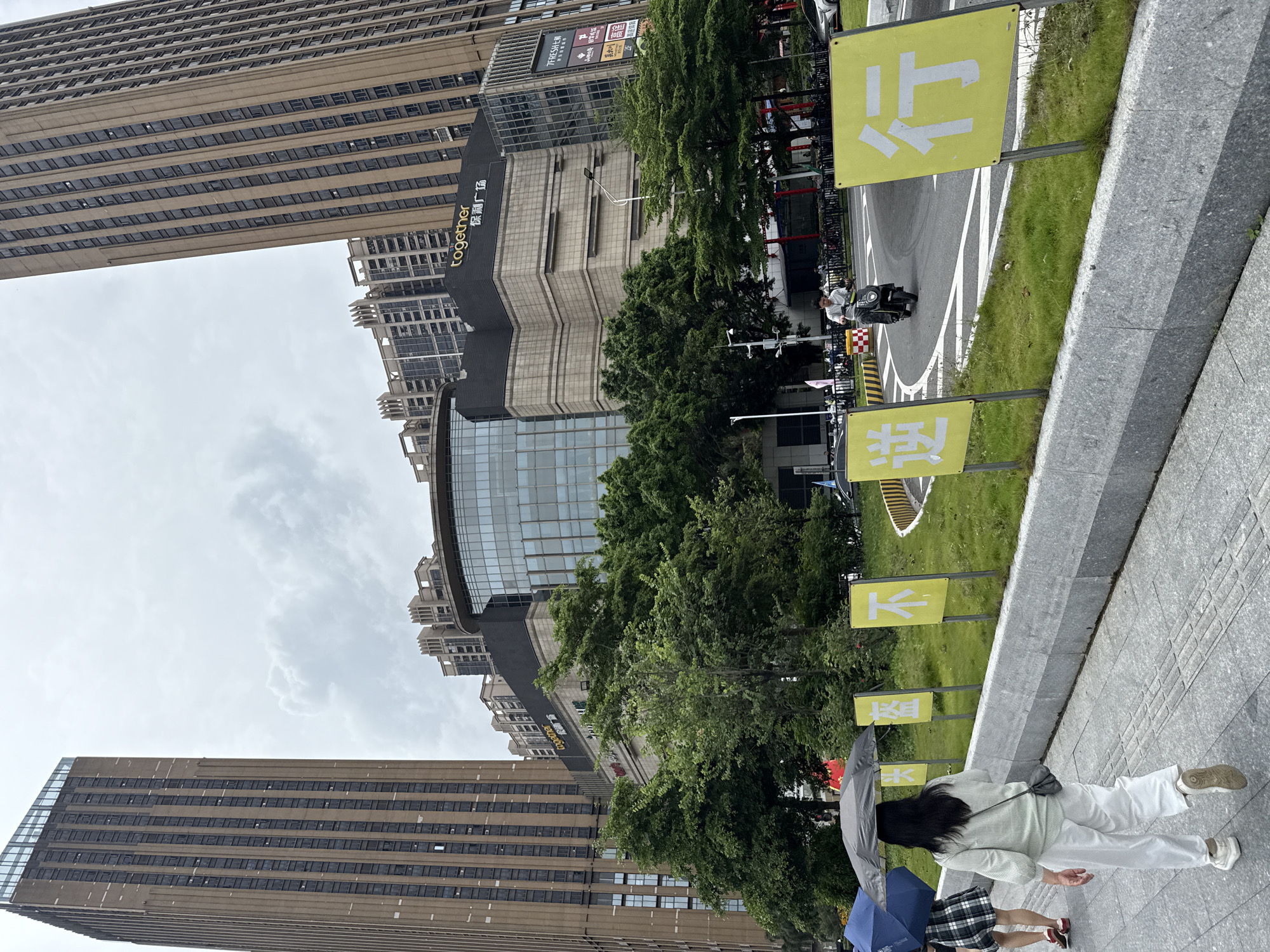 In Haizhu District, China, a street scene unfolds under a cloudy sky. Two towering, modern high-rise buildings, one on the left and one on the right, frame a large, multi-story shopping complex in the background. The complex features a curved glass facade and a dark upper section with a prominent "together" logo and Chinese characters. Trees and greenery are visible in front of the complex.
In the foreground, a sidewalk runs along a grey stone curb. Two women are walking on the sidewalk, one further ahead and to the left holding a blue umbrella, and another closer and to the right, holding a grey umbrella. Both are dressed casually, with the woman closer to the camera wearing white pants and a light-colored top. A person on a scooter rides on the street in the middle ground, to the right of the women. Several yellow signs with white Chinese characters are planted in the grass along the curb, facing the street. The sign closest to the viewer displays the character "行" (xíng, meaning to go or to travel). Further to the left, other yellow signs are partially visible. The street has white lane markings and a curved section. In the far background, more apartment buildings are visible.