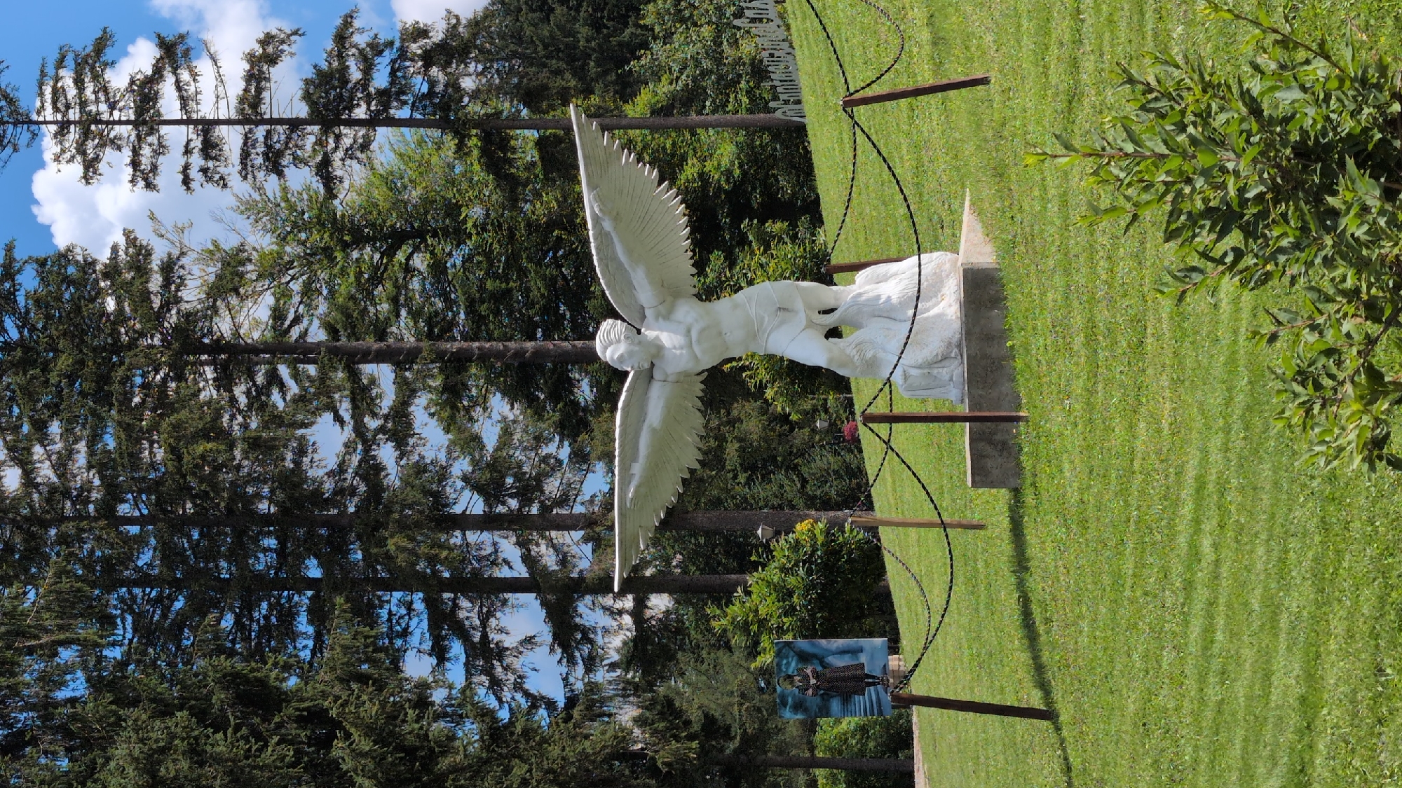 The image shows a white sculpture of a winged figure standing on a rock formation, with its arms outstretched. The sculpture is located in a grassy area with trees in the background. There is a fence made of chains and wooden posts surrounding the sculpture. To the left of the sculpture, there is a large vertical poster or artwork depicting a woman in a patterned dress. The environment appears to be an outdoor park or garden. The sky is blue with some white clouds. In the background, on a grassy hill, is a white sign that reads "Cantacuzino Castle." This indicates the location is likely near Cantacuzino Castle in Bușteni, Romania, which is known for its outdoor sculptures and gardens.