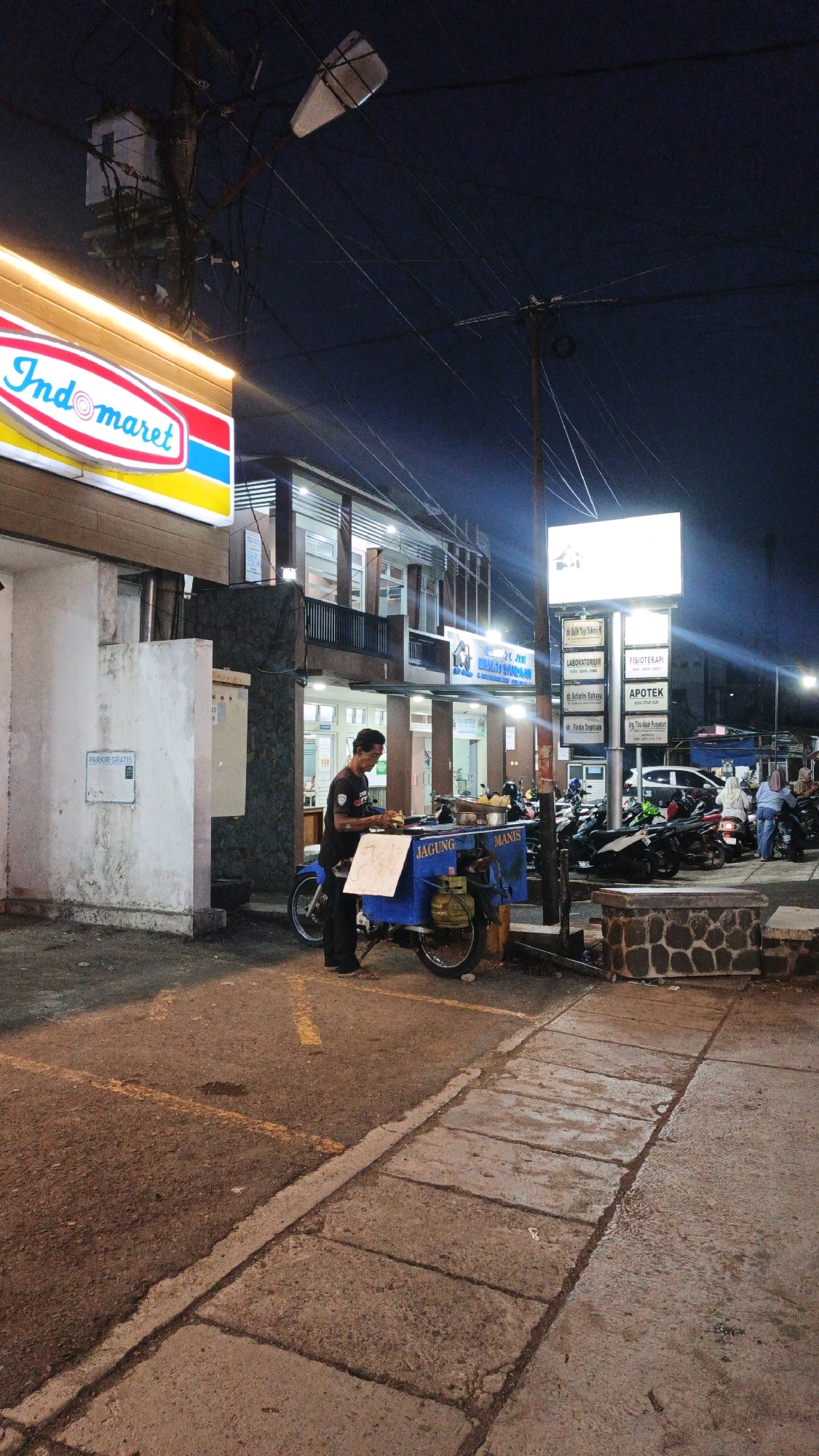 This image depicts a nighttime scene outside an Indomaret convenience store in Cibenda, Indonesia. A man is standing next to a blue mobile food cart, which has the words "JAGUNG MANIS" (sweet corn) written on it. He appears to be preparing food. The food cart is attached to a motorcycle. In the background, there is a building with multiple signs, including one for an "APOTEK" (pharmacy) and others listing medical services. Several motorcycles are parked in front of the building, and some people are visible walking or riding them. The scene is illuminated by streetlights and the lights from the store and signs. The ground is paved with asphalt and concrete.