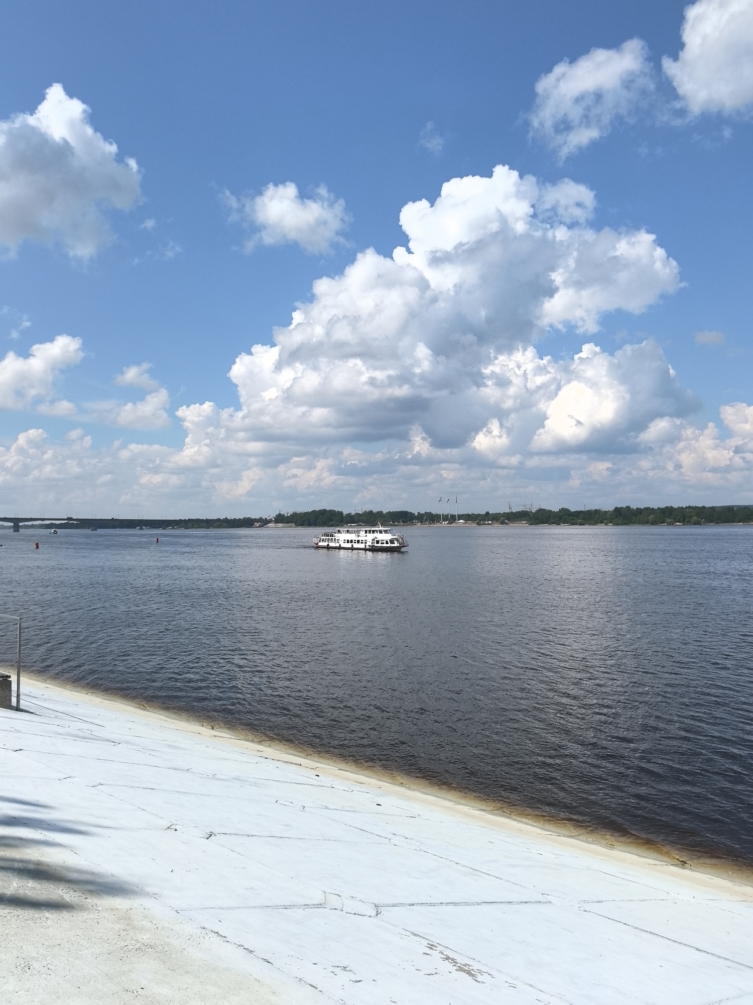 An outdoor, eye-level, vertical, medium shot captures a wide, dark blue river under a bright blue sky filled with fluffy white clouds. A white passenger boat with multiple windows and decks is sailing on the right side of the river. The riverbank on the left is a paved, white embankment sloping down to the water. In the distance, a long, dark bridge spans the river, with trees lining the far bank. The scene appears to be the Kama River in Perm, Russia, likely near the city's embankment.