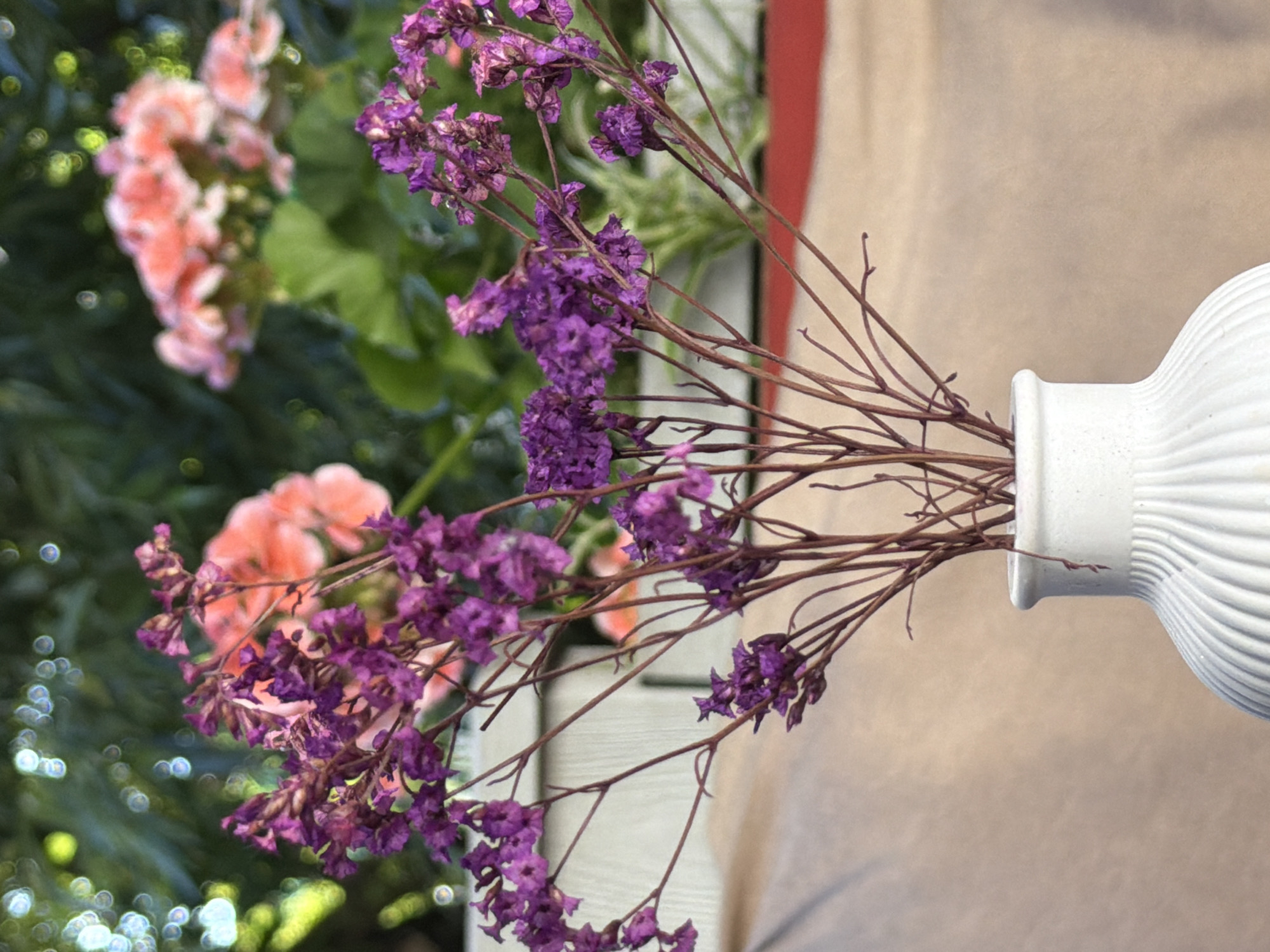 A close-up, eye-level shot focuses on a white, ribbed ceramic vase holding a bouquet of dried purple flowers. The vase is situated on a surface covered with a beige cloth, which is partially obscured by a red object in the lower right quadrant. The background is softly blurred, revealing lush green foliage, delicate pink flowers, and the bokeh effect of distant lights. The overall setting appears to be an outdoor garden or patio. There are no people or animals visible in the image.