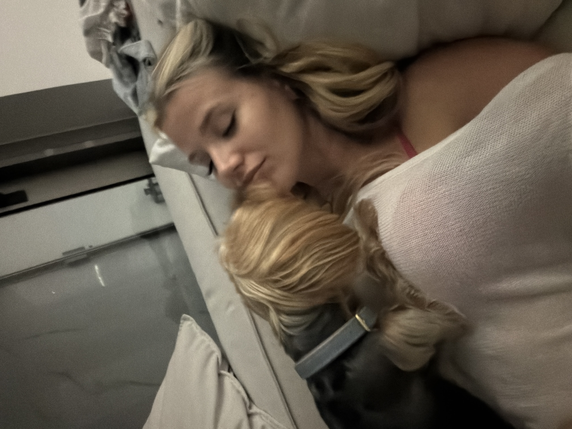 A young woman with blonde hair lies on a couch, her eyes closed. A small, tan-colored dog is leaning in towards her face, as if kissing or nuzzling her. The woman is wearing a light-colored garment, possibly a sweater or blanket, over a pink top. The environment appears to be indoors, with a couch and pillows visible. There are no specific elements in the image that would indicate the location in Thep Krasatti, Thailand.