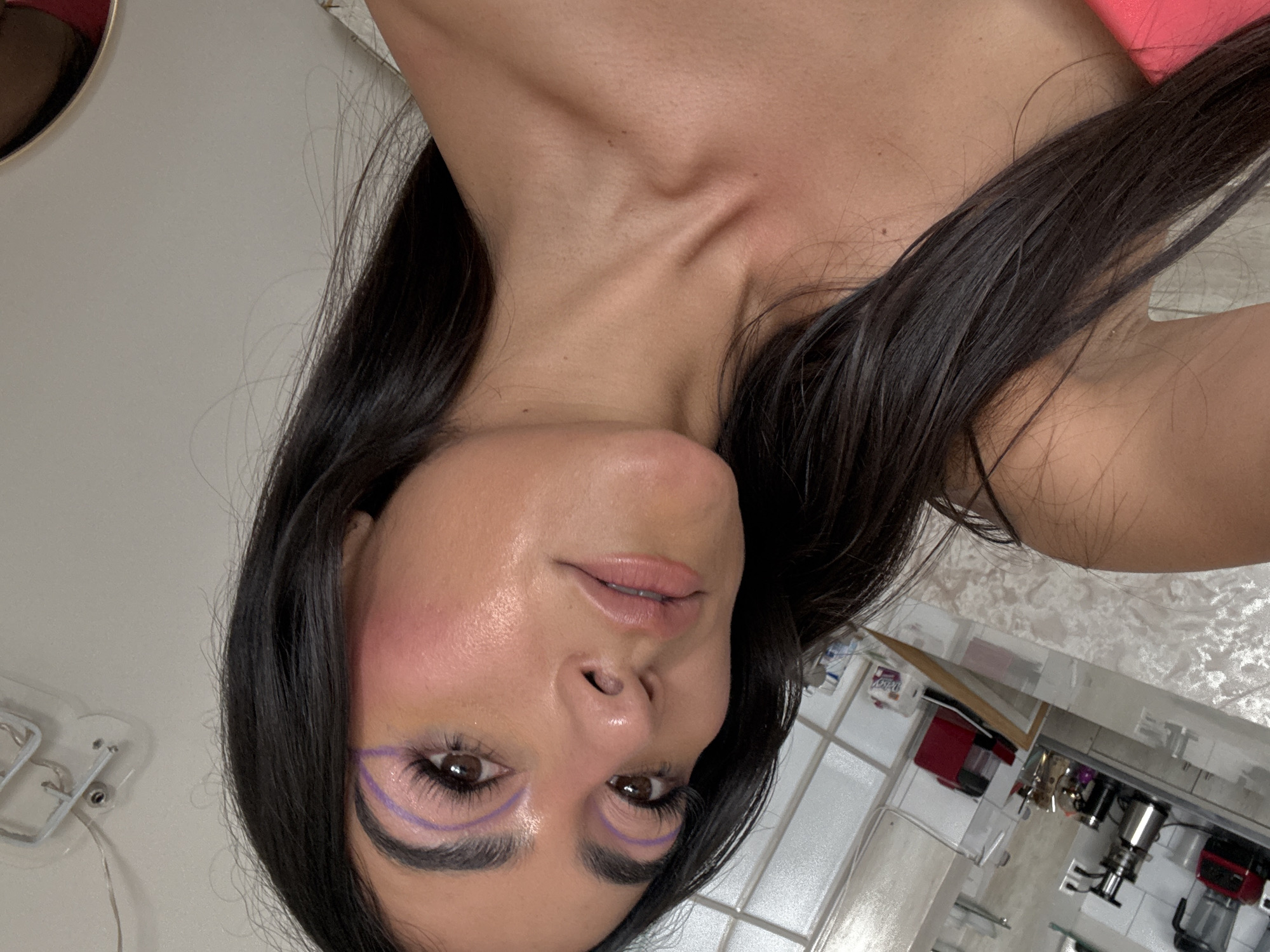 A woman with long, dark hair is pictured from a slightly tilted, close-up angle. She is wearing a subtle makeup look with a focus on her eyes, which feature a purple, arched eyeliner detail. Her skin appears smooth and has a slight sheen. The background includes a glimpse of a kitchen area with white tiled walls, various kitchen appliances (including a blender and a coffee machine), and a marble countertop. The image was taken in Puebla, Mexico.