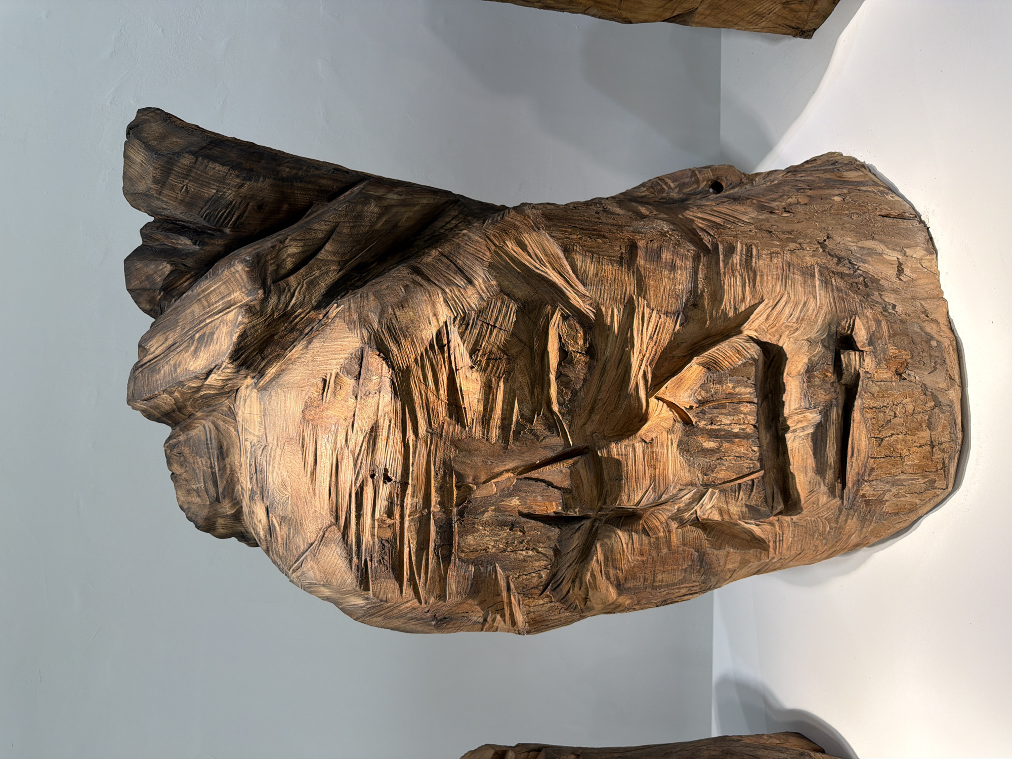 A large, intricately carved wooden sculpture resembling a human face is displayed on a white surface against a plain grey wall. The sculpture is positioned centrally in the frame and is lit from the side, highlighting the deep chisel marks and the natural grain of the wood. The face is stylized, with hollowed-out eyes and a gaping mouth, giving it a primitive or expressive quality. The texture of the wood is rough and weathered, with variations in color from dark brown to lighter golden hues. The background is stark, with the grey wall subtly textured and illuminated. To the left and right of the central sculpture, parts of other similar wooden sculptures are visible, suggesting they are part of an exhibition or collection. The scene is static and appears to be an indoor display, possibly in a gallery or museum. The image is a high-resolution, well-lit photograph that captures the detail and character of the artwork. The location of this artwork is Chongqing High-tech Zone, China, specifically at the "The Mountain's Soul" art exhibition at the Chongqing Art Museum, an institution dedicated to showcasing diverse artistic expressions and cultural heritage within the region.