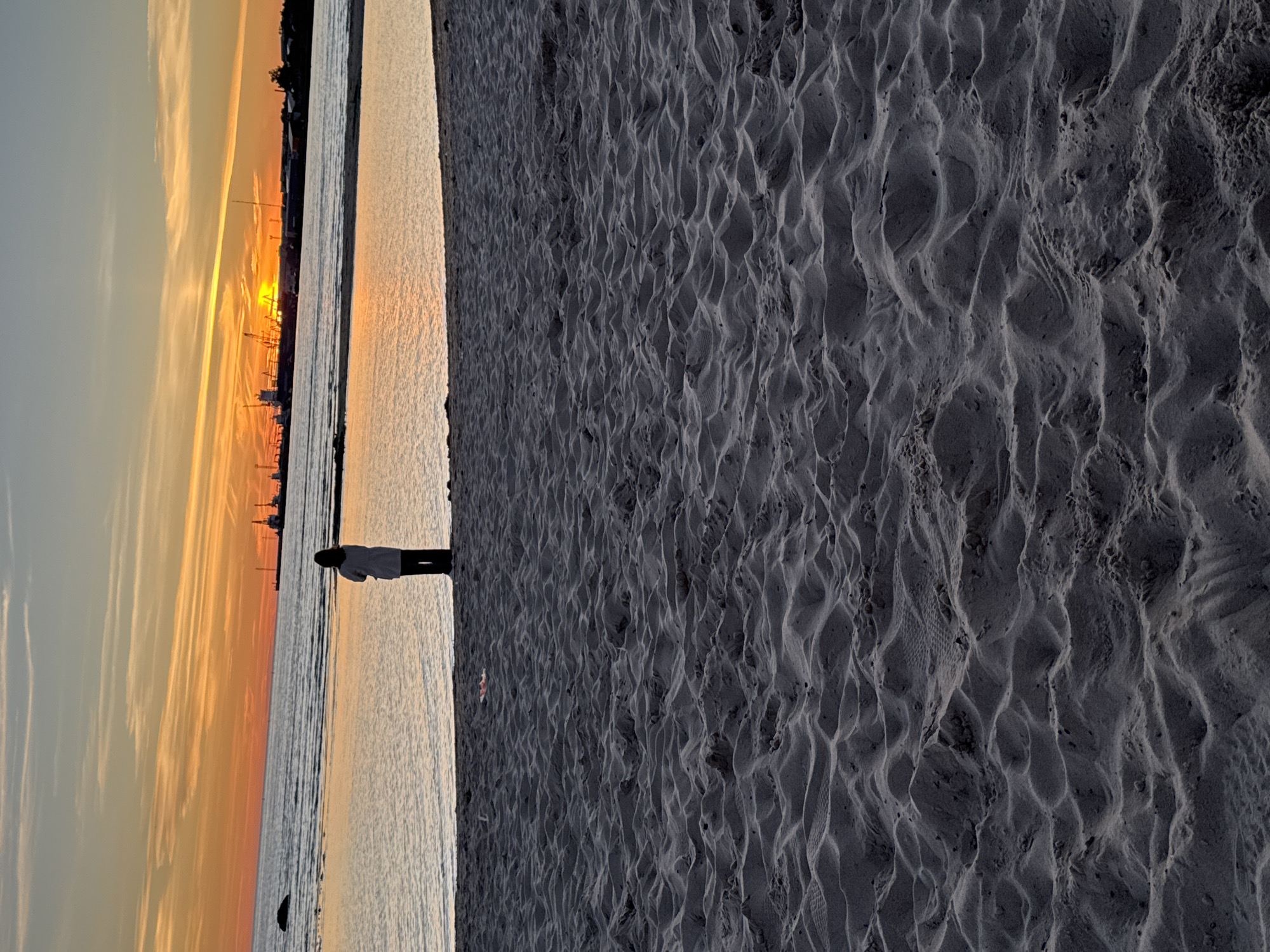 At sunset on a beach in Tallinn, Estonia, a lone figure stands facing the water. The sky is a vibrant canvas of orange and yellow, with streaks of pink and purple clouds. The sun, a bright orb, dips below the horizon, casting a warm glow on the scene. The calm sea reflects the colors of the sky, creating a mirror-like effect. In the distance, industrial cranes and structures of the harbor are silhouetted against the vibrant sky. The foreground is dominated by the textured surface of the sandy beach, with subtle ripples and undulations that hint at recent tides. The overall atmosphere is one of tranquility and contemplation, with the solitary figure a small presence against the vastness of the natural elements and the urban backdrop.