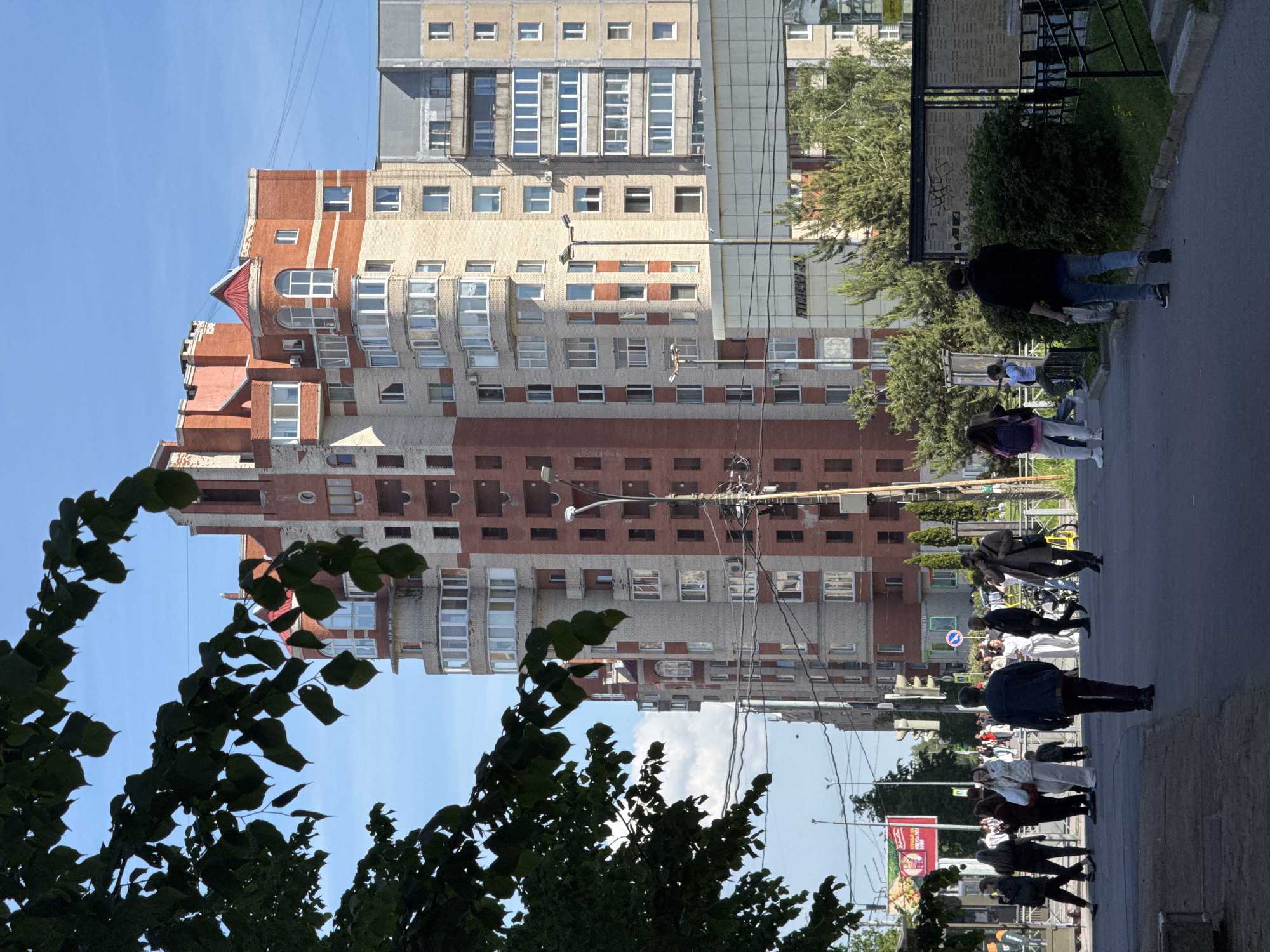The image captures an urban street scene in Saint Petersburg, Russia, on a bright, sunny day. The dominant subject is a tall, multi-story residential building with a varied facade, featuring sections of red brick, lighter beige brick, and grey panels. The architecture showcases a mix of styles, including arched windows, projecting balconies, and a distinctive red-roofed turret-like element at the top. Numerous windows are visible across the building's surface.
In the foreground, lush green foliage from trees and bushes partially frames the view, with dark silhouettes of leaves against the clear blue sky. A paved sidewalk or roadway extends from the lower right to the middle left of the frame, populated by several individuals. People are seen walking, standing, and interacting on the sidewalk, suggesting a normal day's activity. One person in the immediate foreground wears a black jacket and blue jeans, walking away from the viewer. Other pedestrians are scattered further down the street.
Overhead, a complex network of power lines and a streetlamp are visible against the sky and the building. In the background, beyond the main building, another high-rise structure with a gridded facade can be partially seen.
Notable details include the clear weather with strong sunlight, indicated by bright skies and shadows. Visible text includes "КРАСНОЕ" and "БЕЛОЕ" (Krasnoye & Beloye), likely from a store sign, and "Фитнес 24" (Fitness 24) on a green sign on a building further down the street. A blue circular road sign is also faintly visible on the sidewalk.