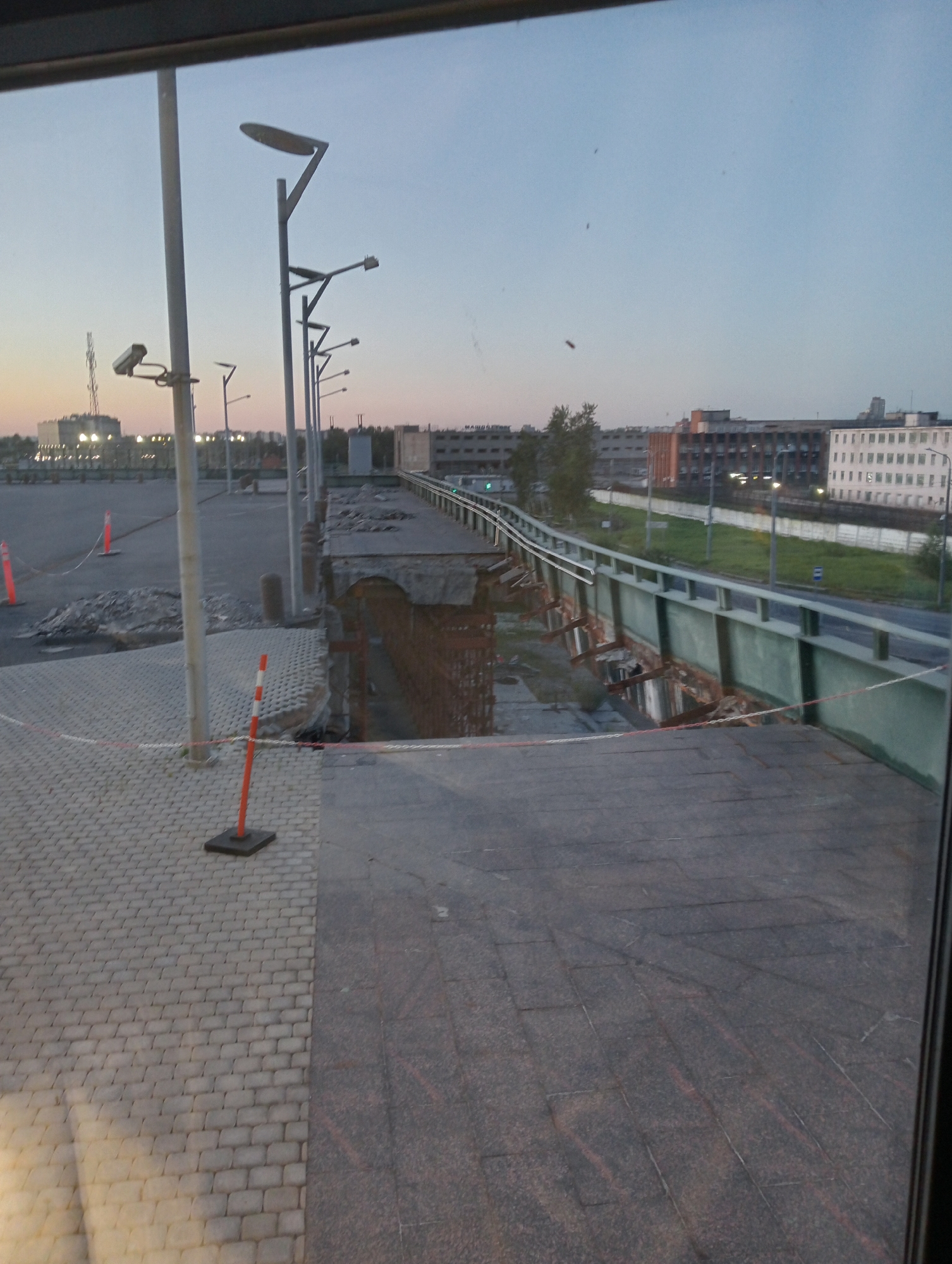 The image depicts an elevated pedestrian walkway that has partially collapsed in Saint Petersburg, Russia. The walkway, constructed of concrete and metal, shows significant structural damage with a large section missing and exposed rebar. A safety barrier, consisting of red and white tape, cordons off the damaged area. Orange traffic cones are also visible, further indicating a restricted or construction zone. The surrounding environment includes paved surfaces, lampposts, and in the background, several multi-story buildings and trees. The sky is overcast, suggesting it is either dawn or dusk. There are no people visible in the image.