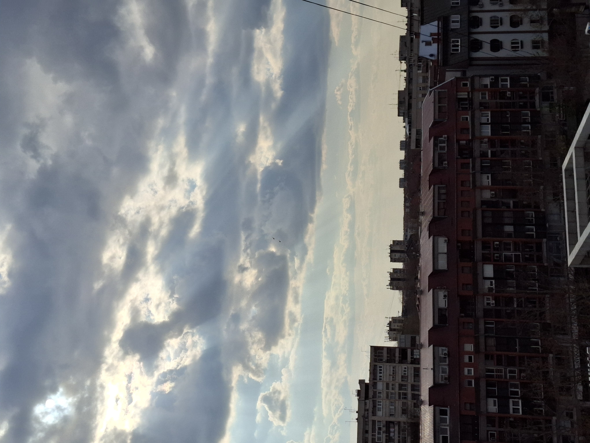 The image depicts an urban landscape in Novi Sad, Serbia. The foreground shows a cloudy sky with visible sunbeams. The background features residential buildings of varying heights and architectural styles, with a mix of red and grey facades. There are several visible windows and balconies. Two birds are flying in the sky. Electrical wires are seen crossing the sky.