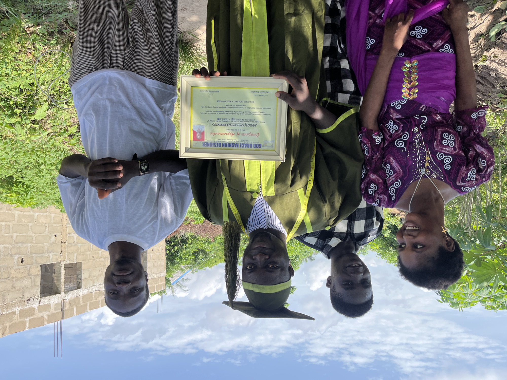 Four people are posing outdoors. The person in the center is wearing a graduation gown with a measuring tape around the neck and holding a certificate that reads "God Grace Fashion Designer, Certificate of Apprenticeship, Adelagun Joshua Opemipo, 3 Years From June 2015 to June 2018, Dated This 16th Day of May Year 2020." The other three people are standing on either side of the central person. The surroundings include vegetation and a partially constructed building. The location is possibly Tofa, Nigeria, based on the certificate text.
