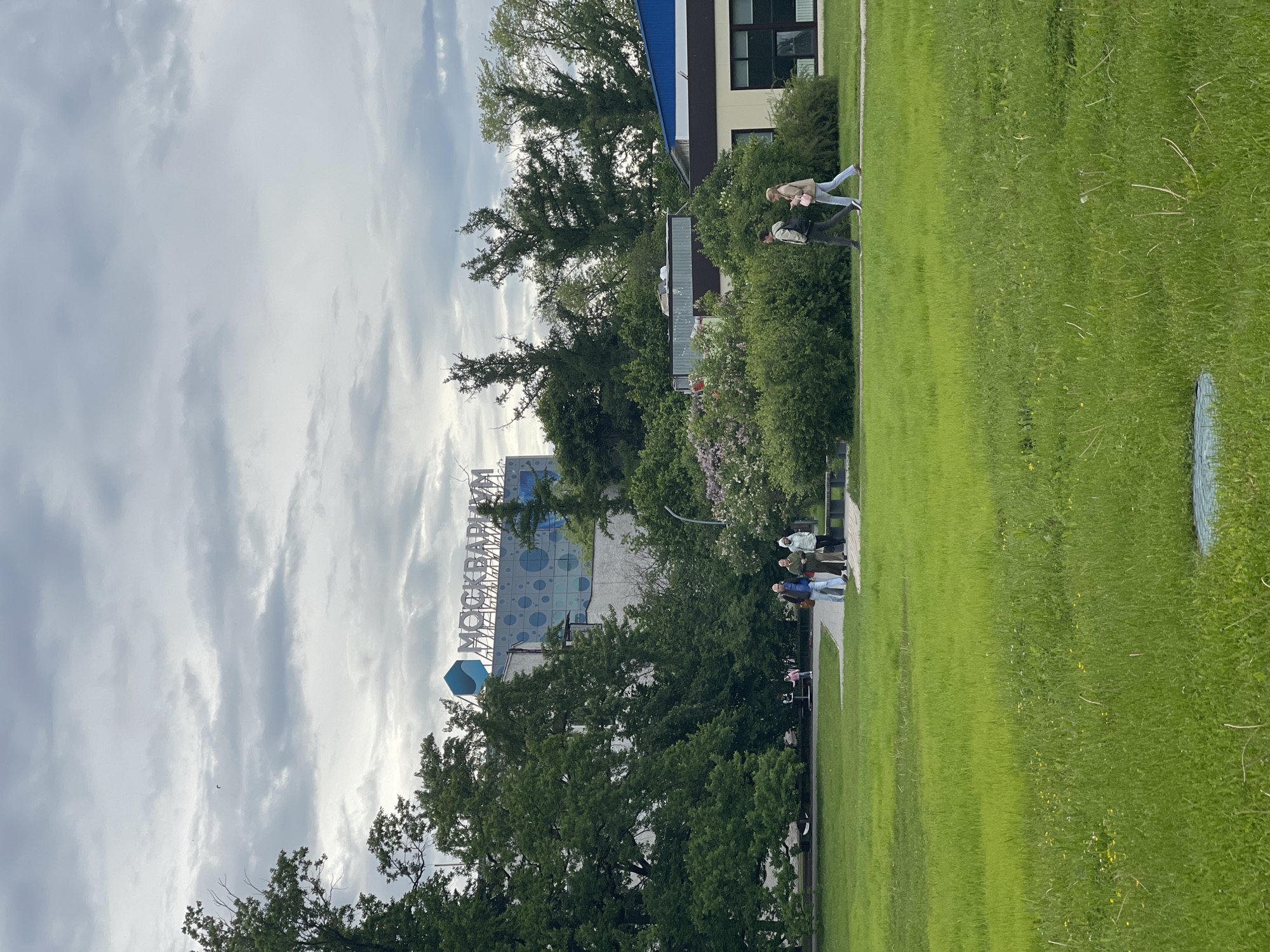The image shows a park or green space. A large building with the word "МОСКВАРИУМ" on top is visible in the background. People are walking or standing along a path in the park. There are trees and bushes in the foreground and middle ground. The sky is cloudy. The location is in Moscow, Russia.