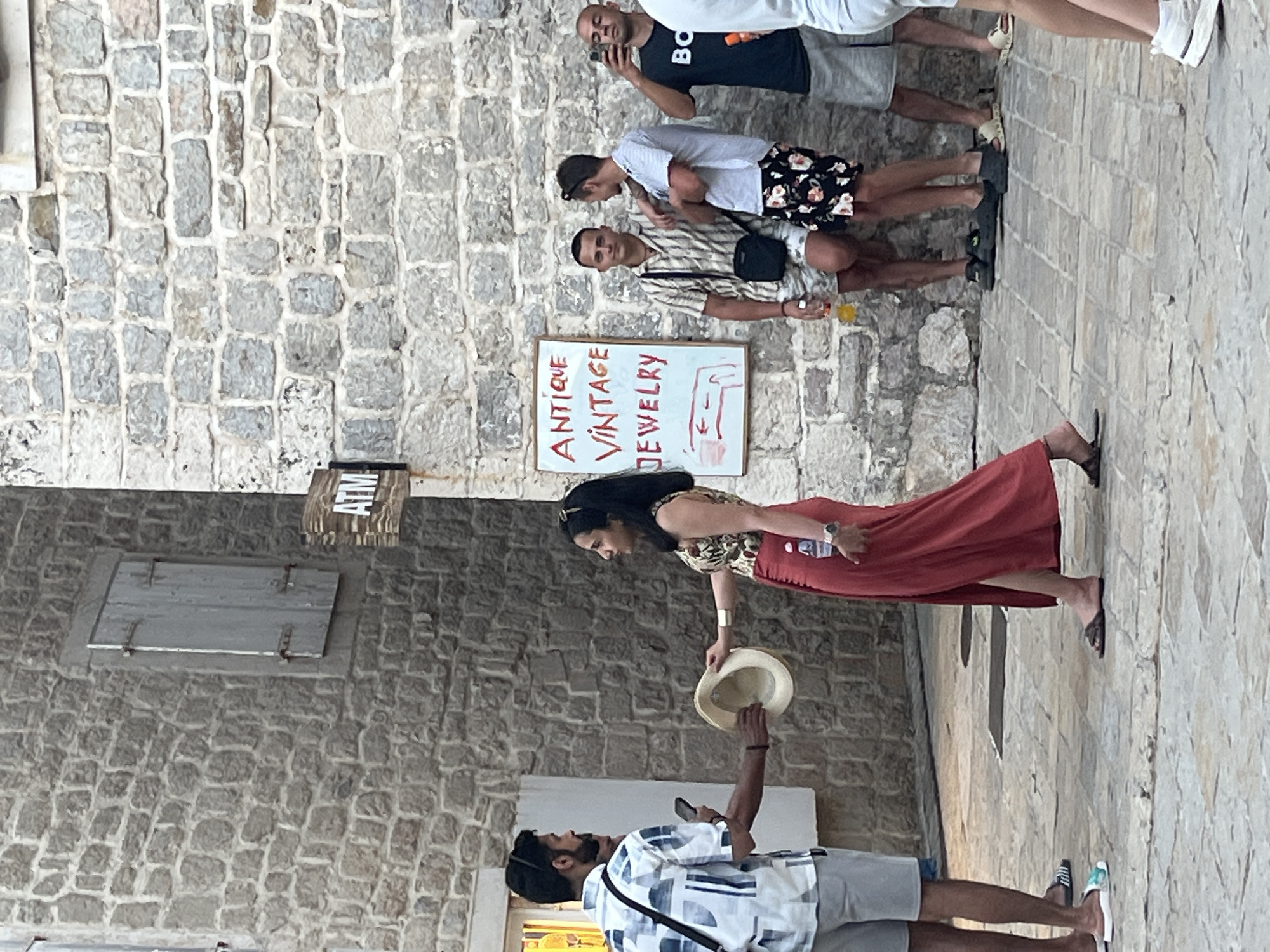The image depicts a street scene in Budva, Montenegro, likely within the Old Town, characterized by its stone buildings. In the foreground, a man in a patterned short-sleeved shirt and grey shorts extends a straw hat to a woman wearing a patterned top and a long red skirt. The woman is holding the hat. Behind them, a white sign with red lettering reads "ANTIQUE VINTAGE JEWELRY" and includes a red drawing of an arm. Several other people are visible in the background: three men standing near the sign, one of whom is holding a phone and appears to be taking a photo, and another man is taking a selfie. The wall behind them is constructed of rough grey stones, and a wooden shuttered window is visible on the left side of the frame. An ATM sign is also present on the building. The ground is paved with stone tiles.