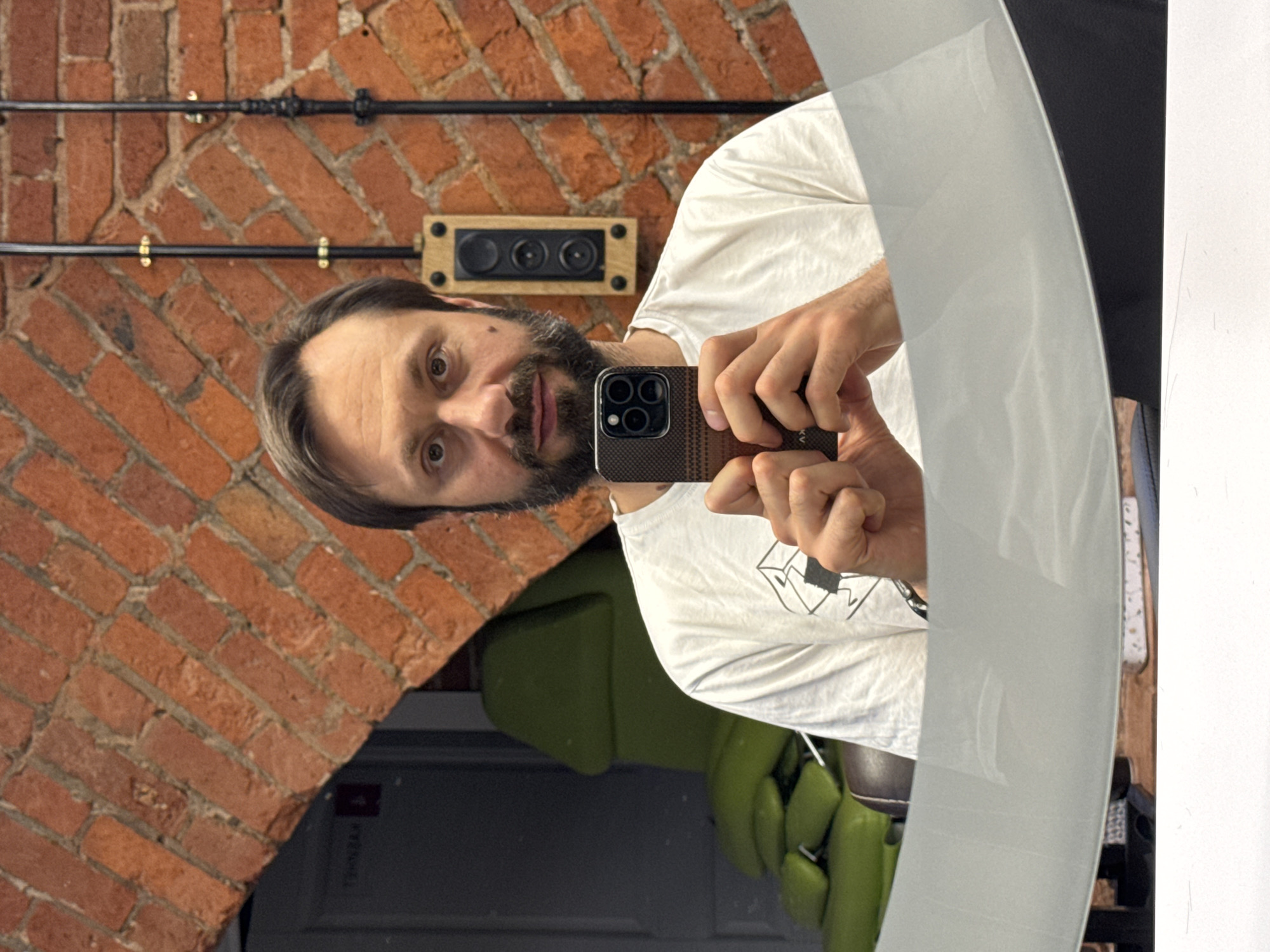 An indoor mirror selfie captures a man in what appears to be a commercial establishment in Moscow, Russia. The man, with a beard and dark hair, is reflected in a large, slightly curved mirror, holding a modern smartphone with a textured dark case (faintly showing "KV" or "K.V." on its corner) with both hands. He is wearing a light-colored t-shirt and looks directly towards the viewer with a neutral expression.
The background reflected in the mirror features an exposed red brick wall, lending an industrial or rustic aesthetic to the space. Black metal pipes or electrical conduits are mounted horizontally across the brick wall, secured by brass-colored clamps. A wooden-framed electrical box or switch plate with three black circular switches or outlets is also visible on the brick surface.
Below the man's arm, a vibrant lime-green, multi-cushioned chair is partially reflected, suggesting it might be a styling or treatment chair, common in barbershops, salons, or tattoo studios. The overall lighting indicates a well-lit interior. A dark area in the lower-left reflection, possibly a doorway, includes a small, unreadable red sign. The image offers a glimpse into a uniquely designed space.