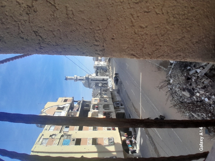The image presents a daytime view of a street in Khan Yunis, Palestinian Territory, framed by two dark vertical bars on the left and a textured, light-brown wall section on the right. In the immediate foreground, a pile of concrete rubble and debris is visible. To the left, a multi-story residential building, approximately six floors high, displays significant structural damage, including exposed interiors, shattered windows, and numerous impact points on its facade. Laundry is visible hanging on balconies of the lower floors.
An asphalt road with white lane markings spans the mid-ground. A single black motorcycle with a rider is observed traveling away from the viewer on the road. Further down the street, a small group of three to four individuals walks near a dark, stationary vehicle. On the far side of the road, a prominent mosque, identifiable by its dome and a tall, slender minaret, stands partially damaged. Surrounding buildings beyond the mosque also show signs of extensive destruction. The sky above is clear blue with scattered faint clouds. The overall scene indicates widespread structural damage across the urban environment. A "Galaxy A14" watermark is visible in the bottom left corner.