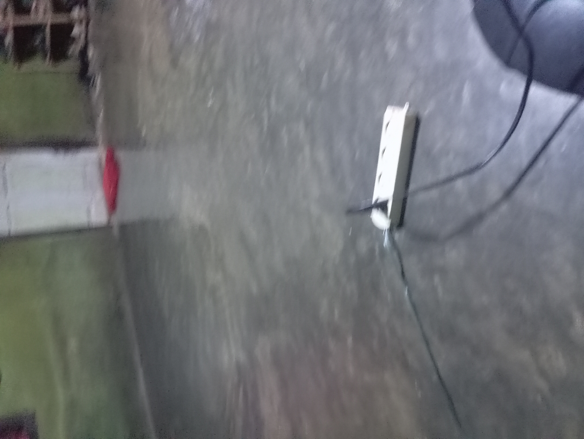 In Rejosari, Indonesia, a room with a concrete floor and greenish-gray walls is depicted. In the foreground, a white power strip with two black cords lies on the floor. One cord extends to the left, disappearing out of frame, while the other curves towards the right, leading to a dark, indistinct object in the lower right corner, possibly a computer mouse. In the background, a red mat is visible on the floor, and a doorway or open passage leads to another area. A shelf unit containing shoes is present on the far right of the image. The scene appears to be an indoor space, possibly a dwelling or utility area.