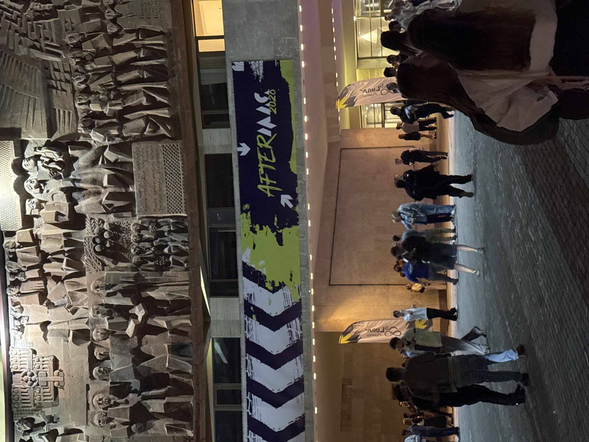 The image shows a group of people standing outside a building in Yerevan, Armenia. On the left is a building with a bas-relief depicting faces. To the right of the relief is a vertical banner with the text "AFTERINS 2026" and arrows. People are standing on a paved surface. Some individuals are blurred. A banner with the image of a bicycle is in the background.