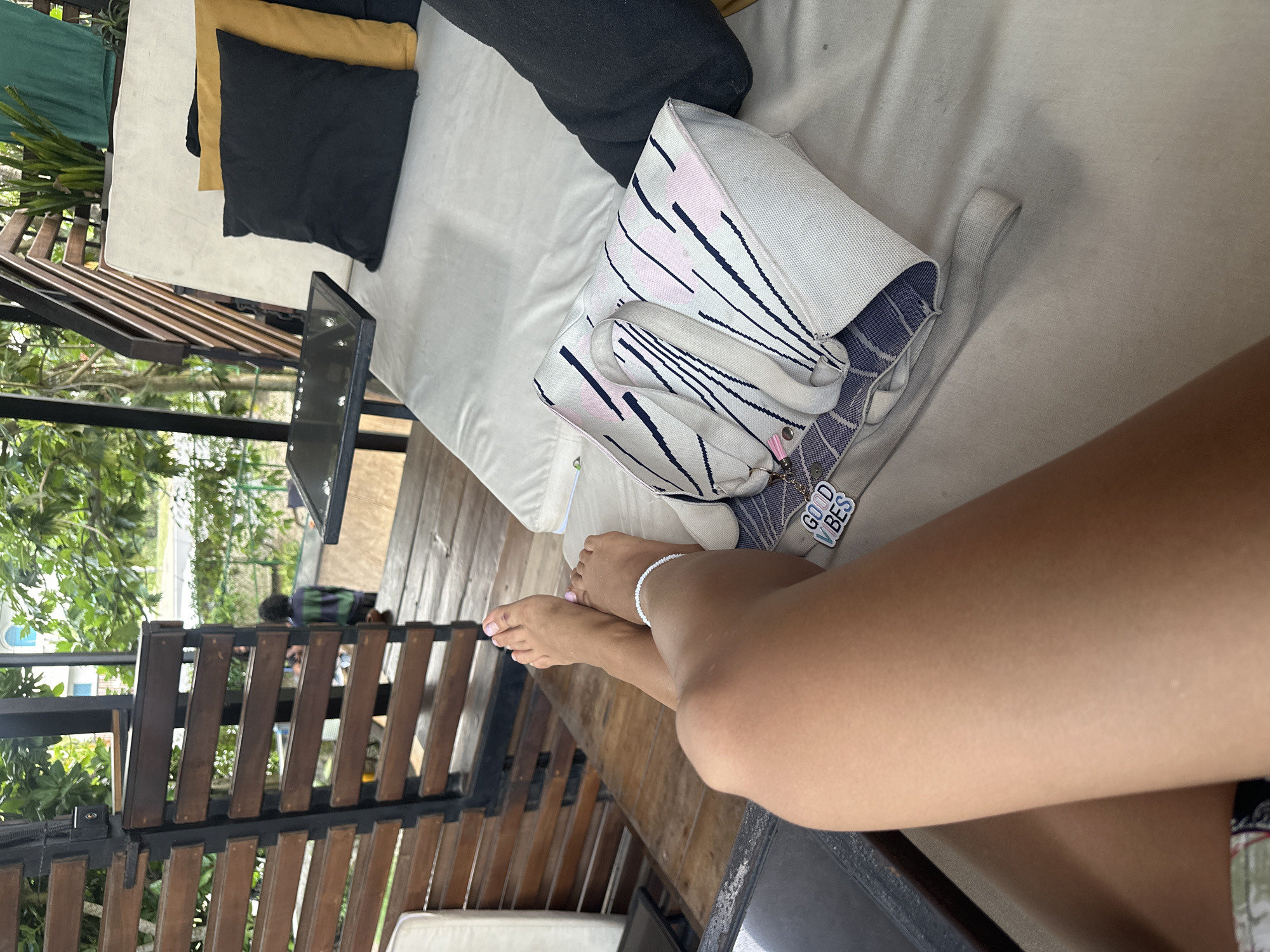The image depicts a scene in Peleana, Sri Lanka. A person's legs are prominent in the foreground, with a white anklet visible on one ankle. The person appears to be relaxing on a cushioned outdoor lounge. A bag rests nearby on the lounge, along with pillows. A laptop sits on a table. The background shows foliage and a wooden railing. Other individuals can be seen in the background.